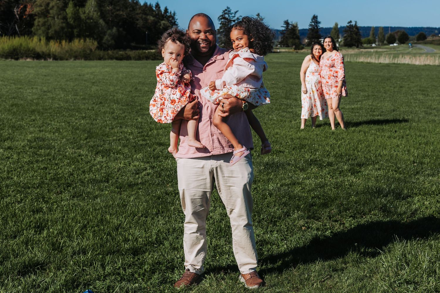 A smiling man holds two young girls in matching floral outfits on a green field, with a family in pink behind him.