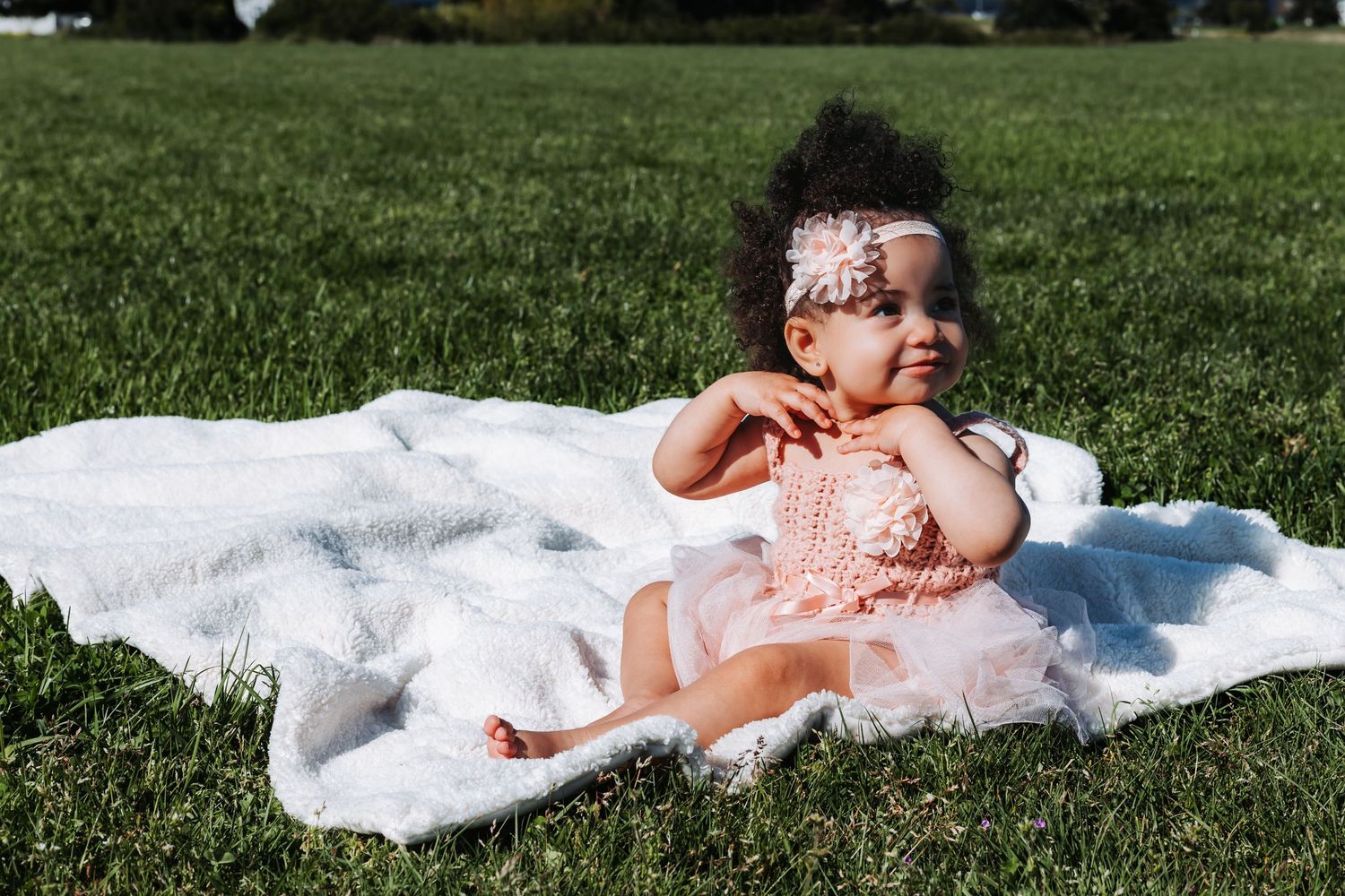 A cute baby girl in a pink dress and floral headband sits on a white blanket on green grass outdoors.