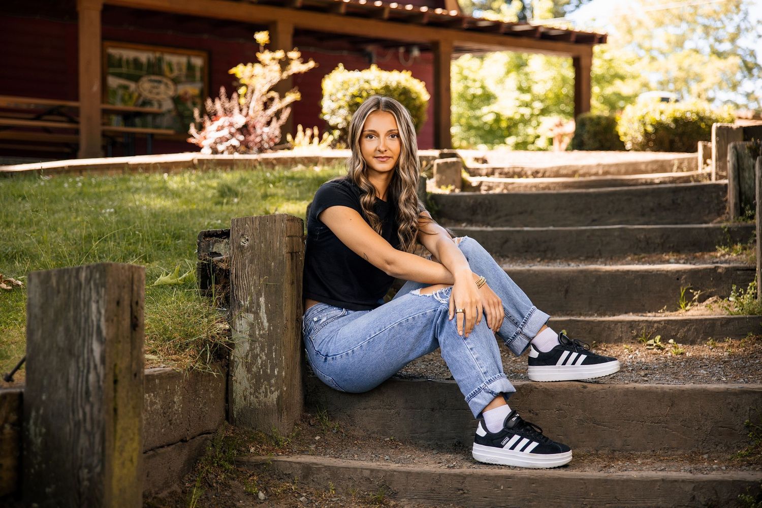 Young woman in black top and jeans sitting on wooden steps outdoors during golden hour.