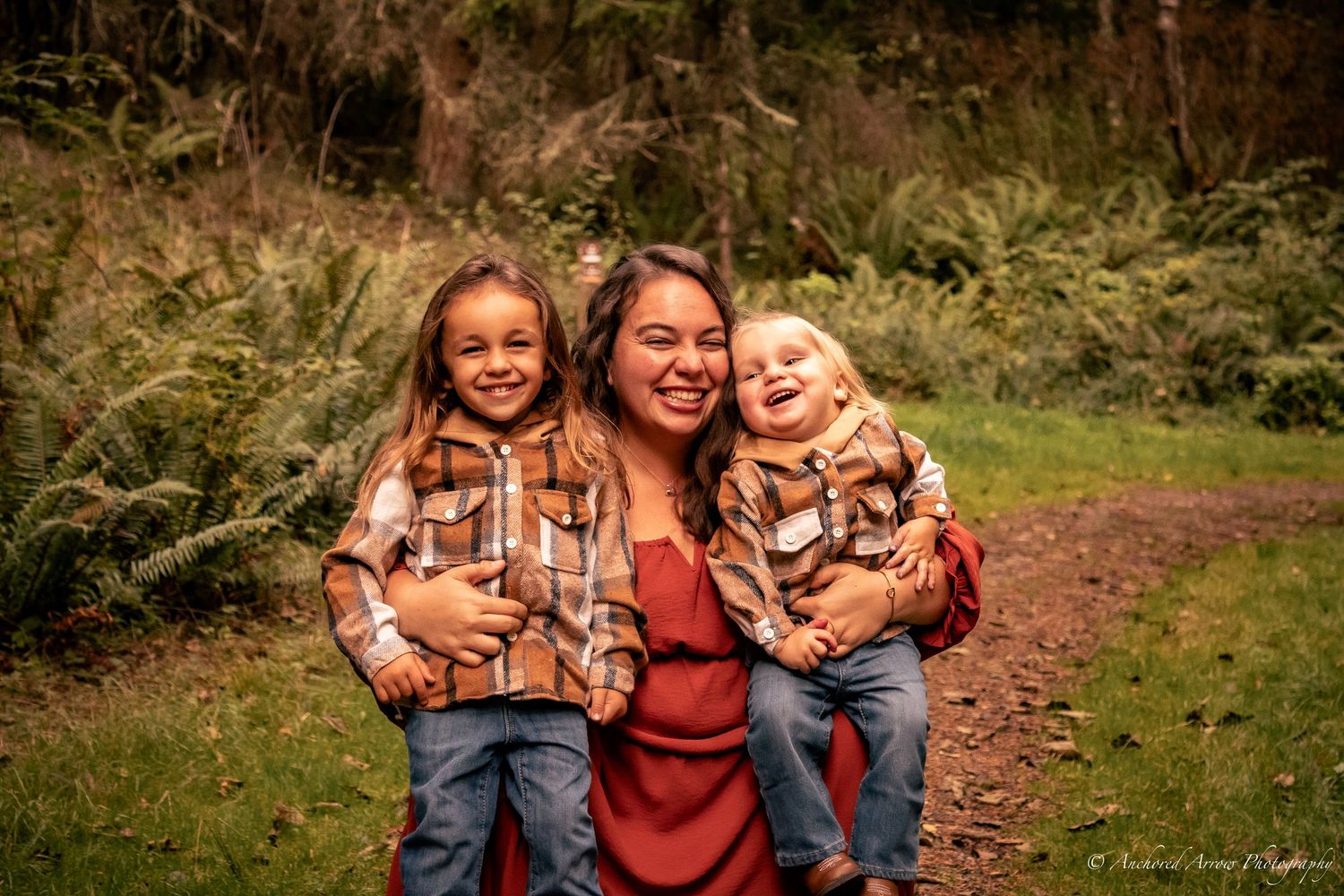 A smiling mother holds two laughing children on a forest path, all wearing plaid shirts and jeans.