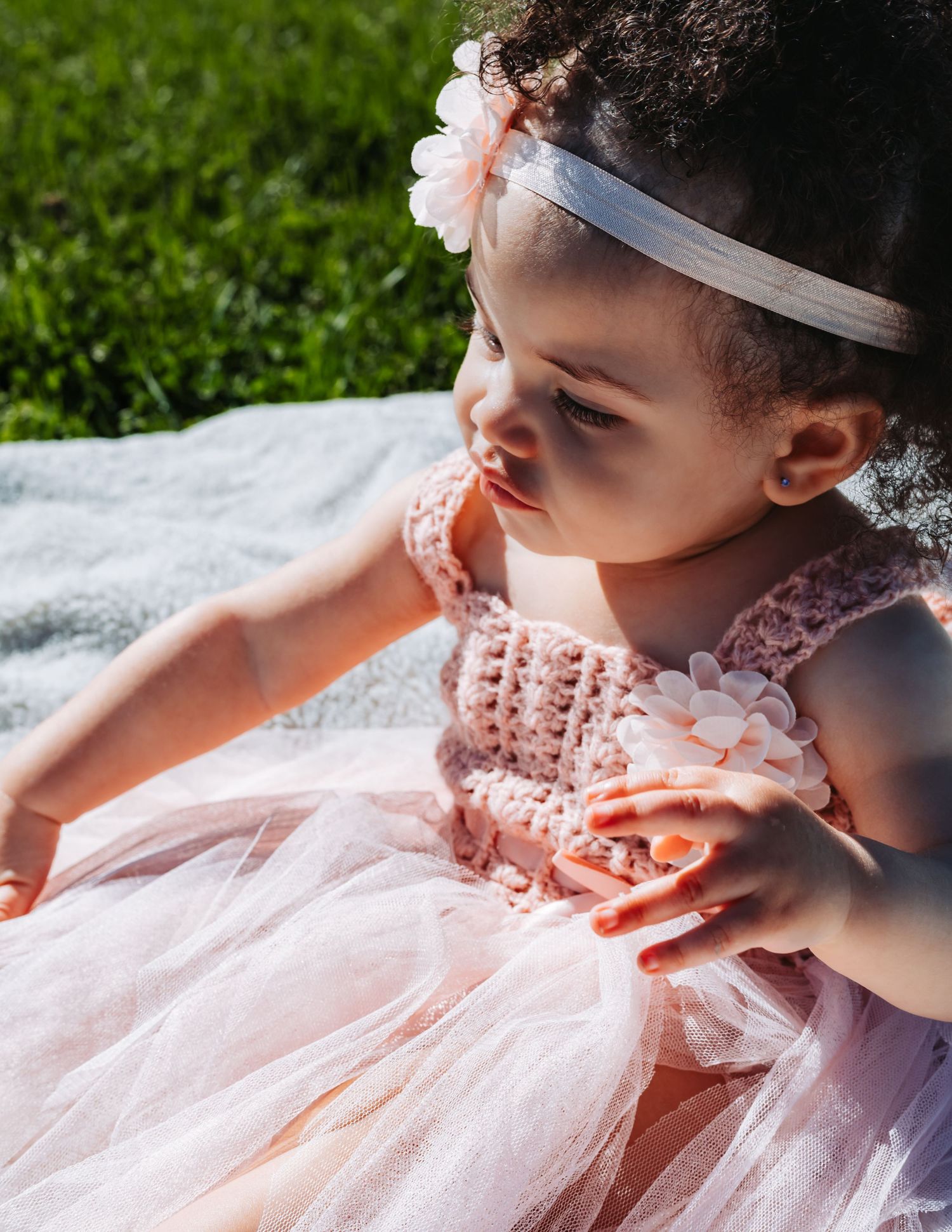 A toddler girl in a pink crochet tutu dress and floral headband sits on a blanket outdoors in sunlight.