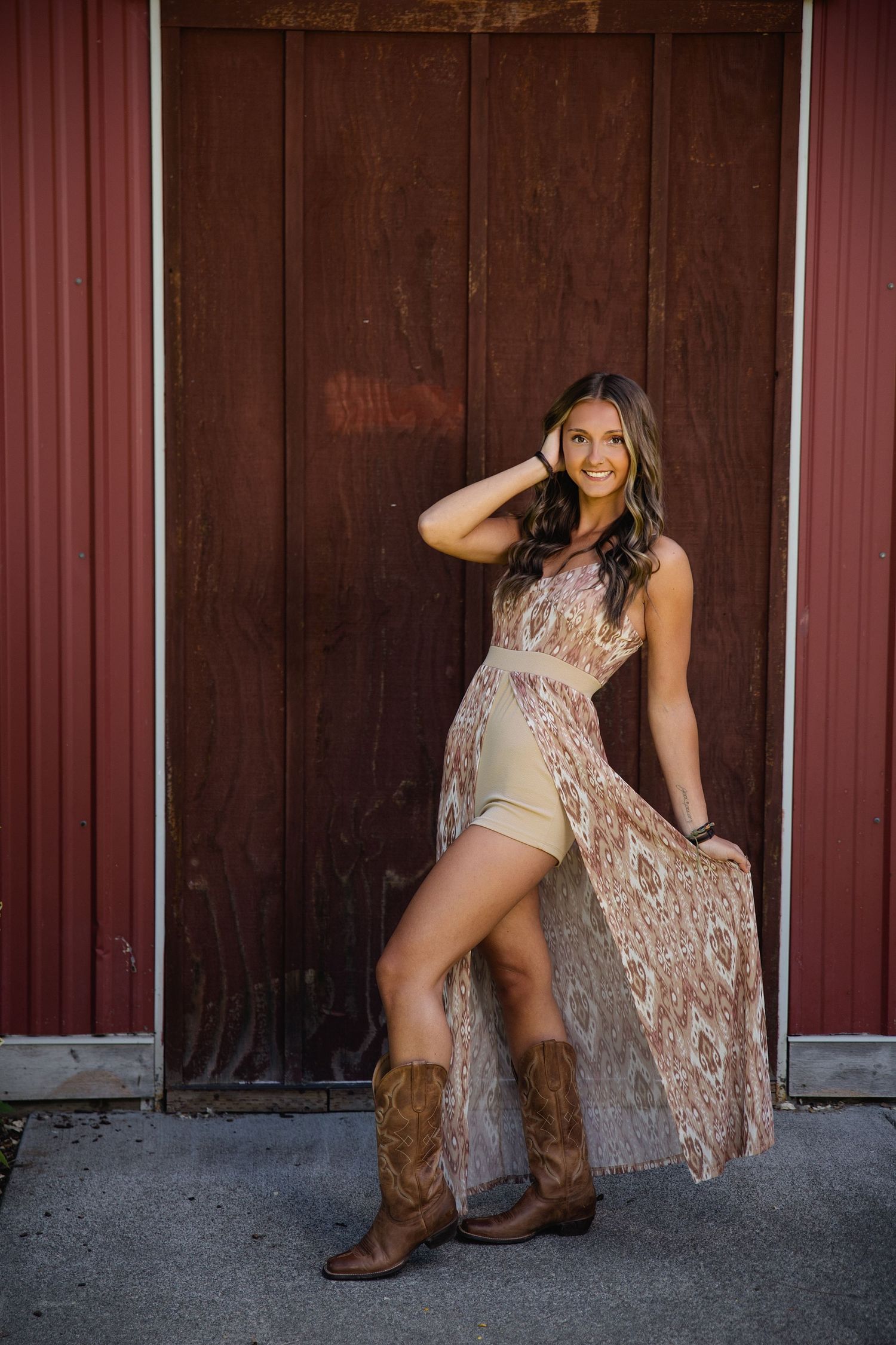Young woman in lace maxi dress and cowboy boots posing confidently in front of a rustic red barn door.