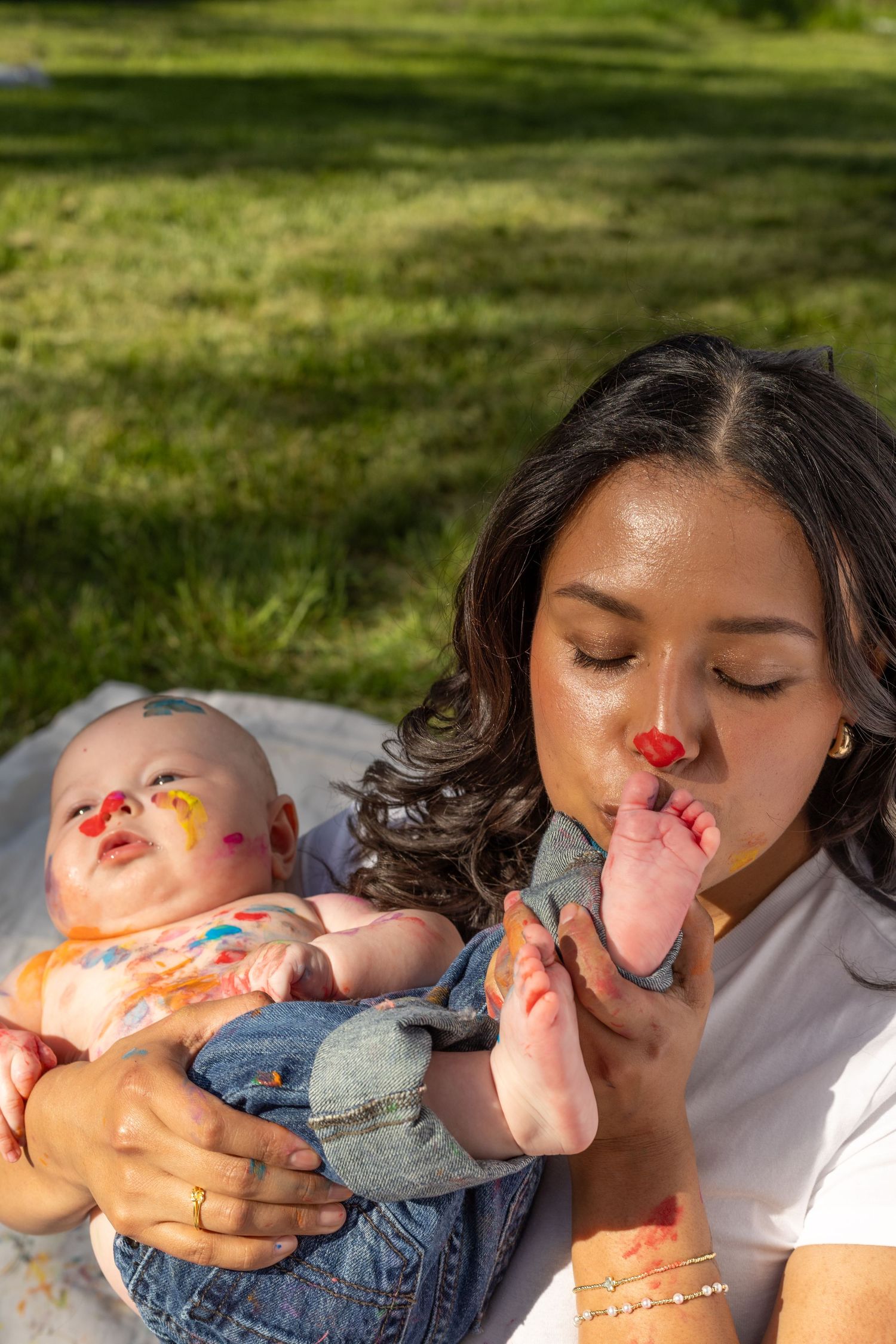 A young mother kisses her baby's painted feet during a colorful outdoor smash cake photoshoot on a sunny day. Mommy and me mini session.