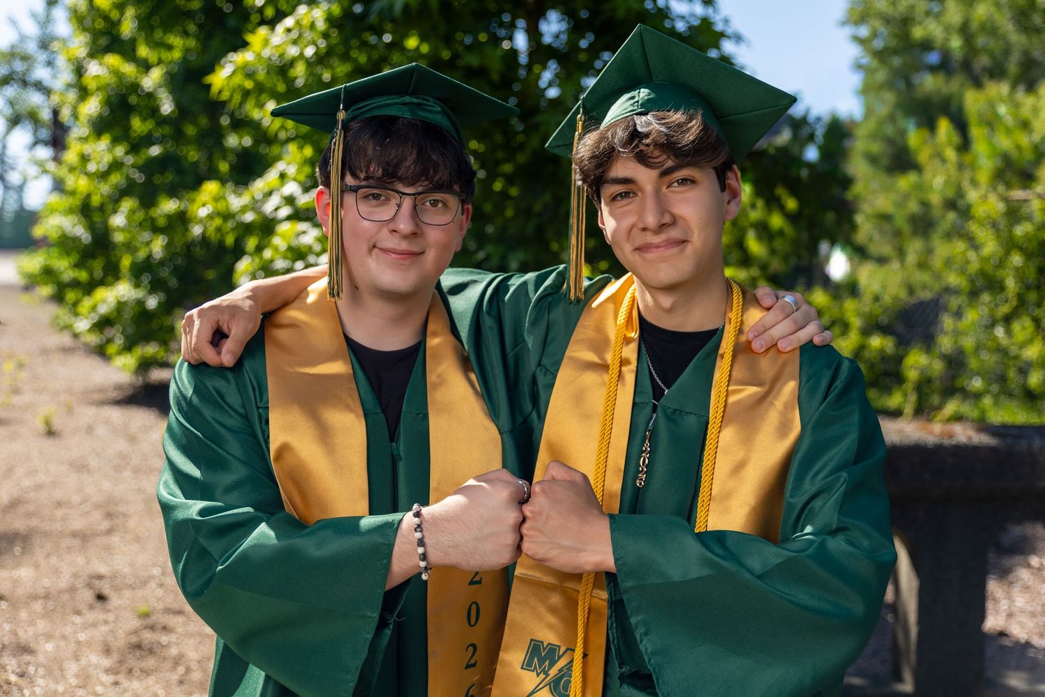 Two male high school graduates in green and gold caps and gowns smiling outdoors.