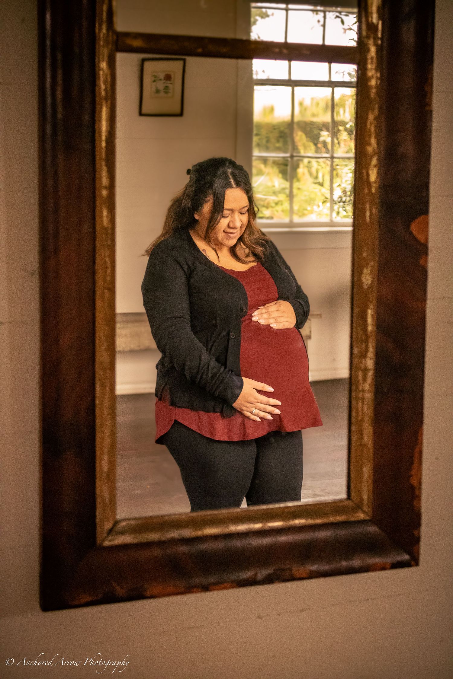 Pregnant woman in red top and black cardigan cradles her baby bump while gazing down, reflected in an ornate wooden mirror.