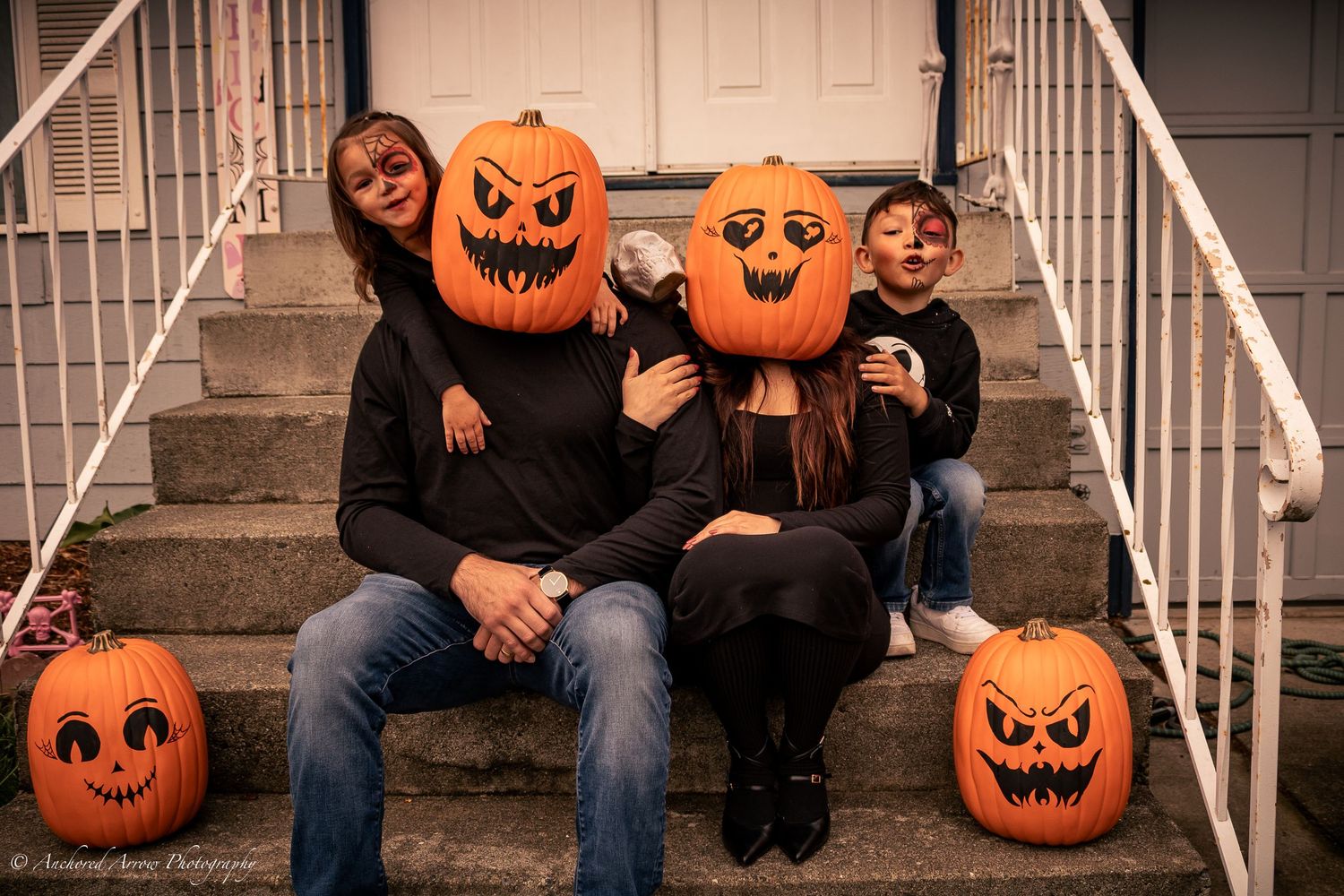 Family posing on steps with carved pumpkin heads for Halloween, surrounded by jack-o-lanterns.