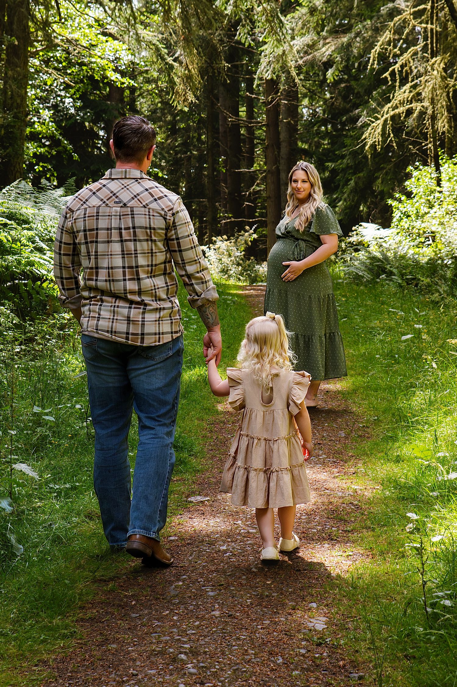 Pregnant woman in green dress stands on forest path as husband and young daughter walk toward her.