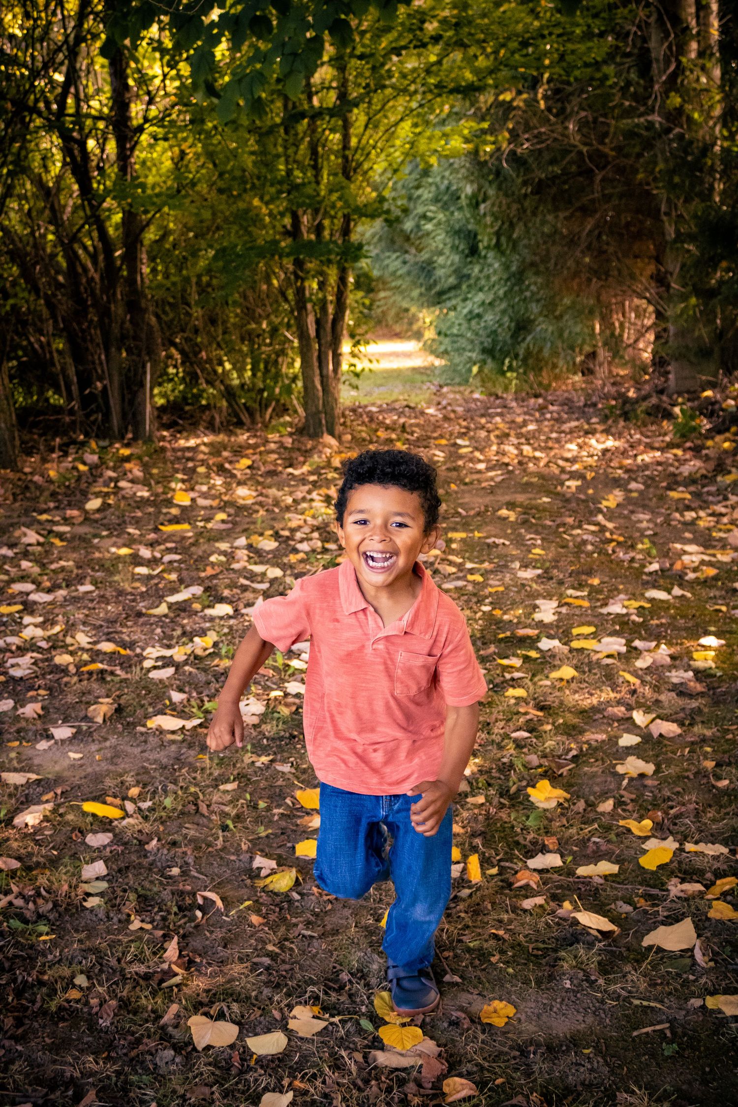 A happy young boy in a coral shirt runs through a leaf-covered autumn forest path.