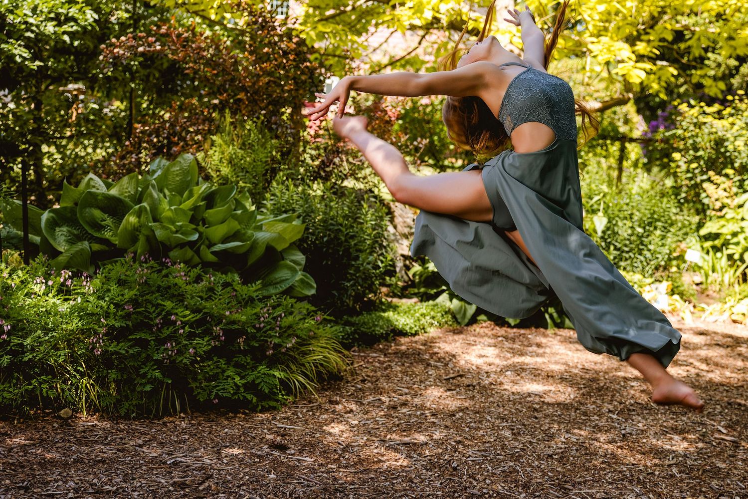 Barefoot dancer in green outfit performs graceful pose among lush garden plants on mulch path.