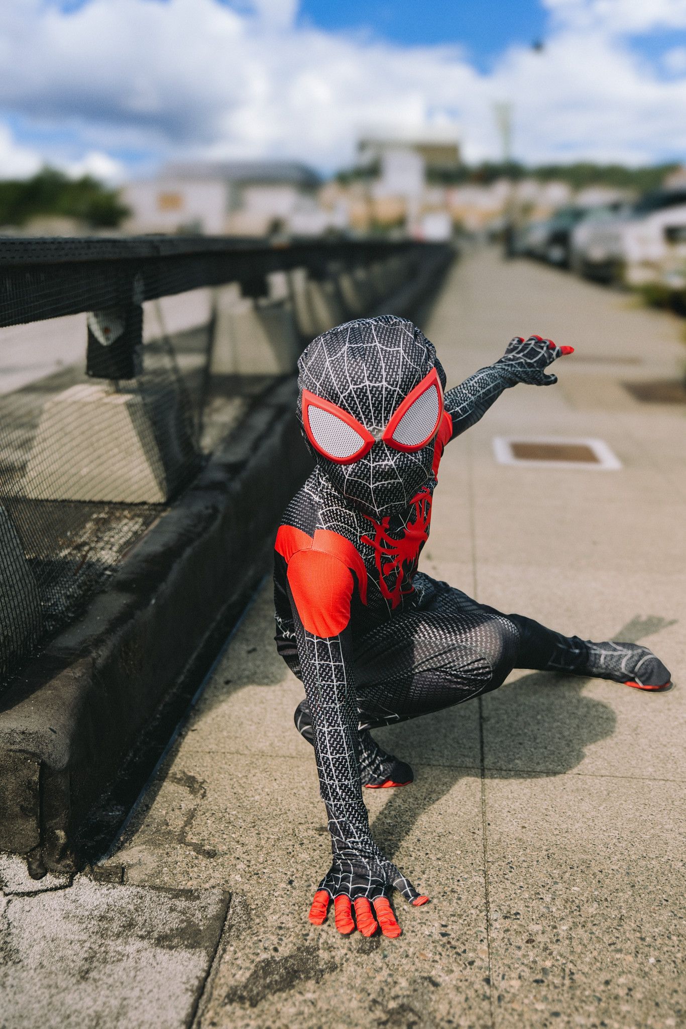 Child in Miles Morales Spider-Man black and red costume poses heroically on a rooftop sidewalk under a cloudy sky.