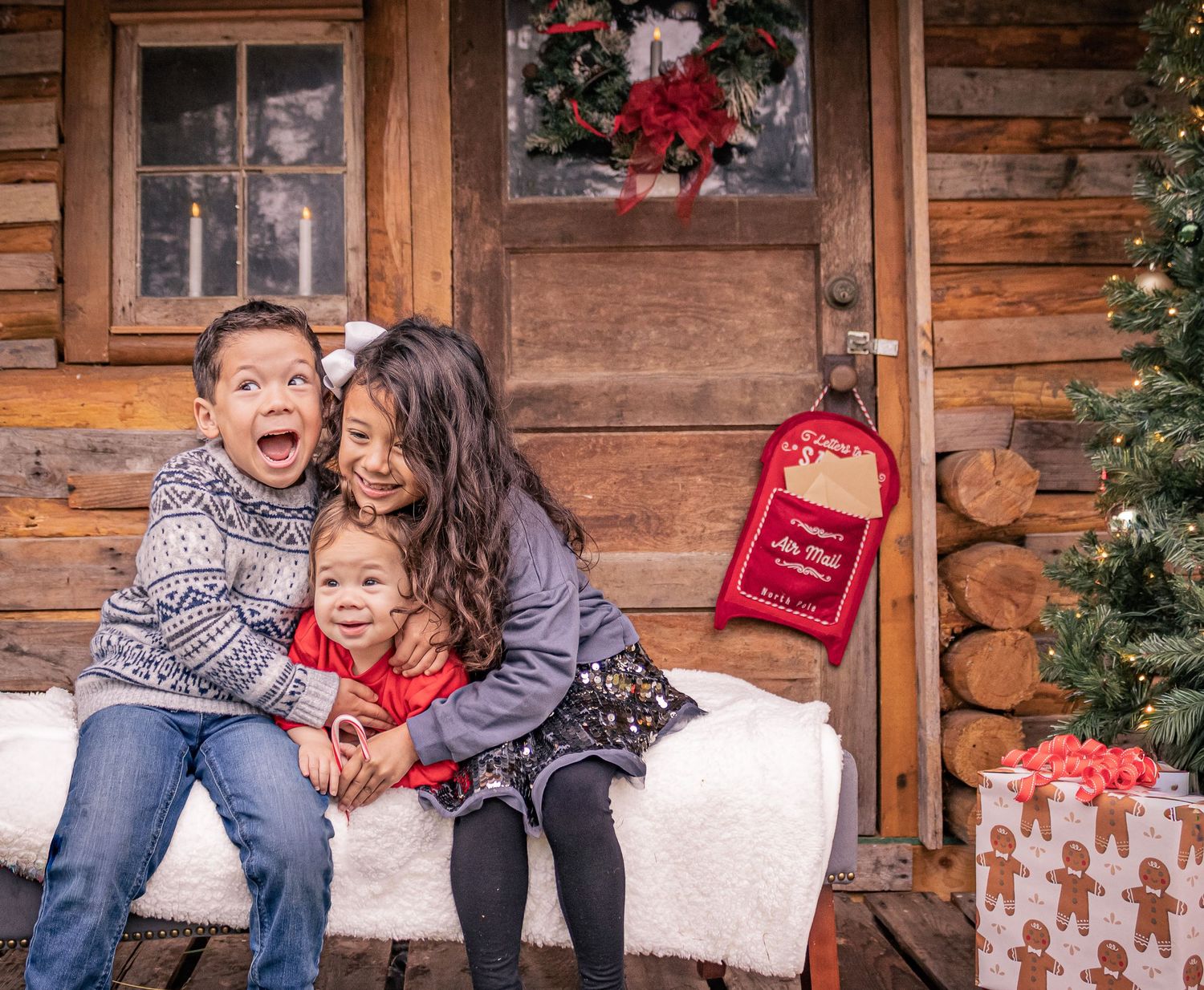 Three children pose joyfully on a snowy bench outside a rustic log cabin decorated for Christmas with a wreath and gifts. Christmas minis