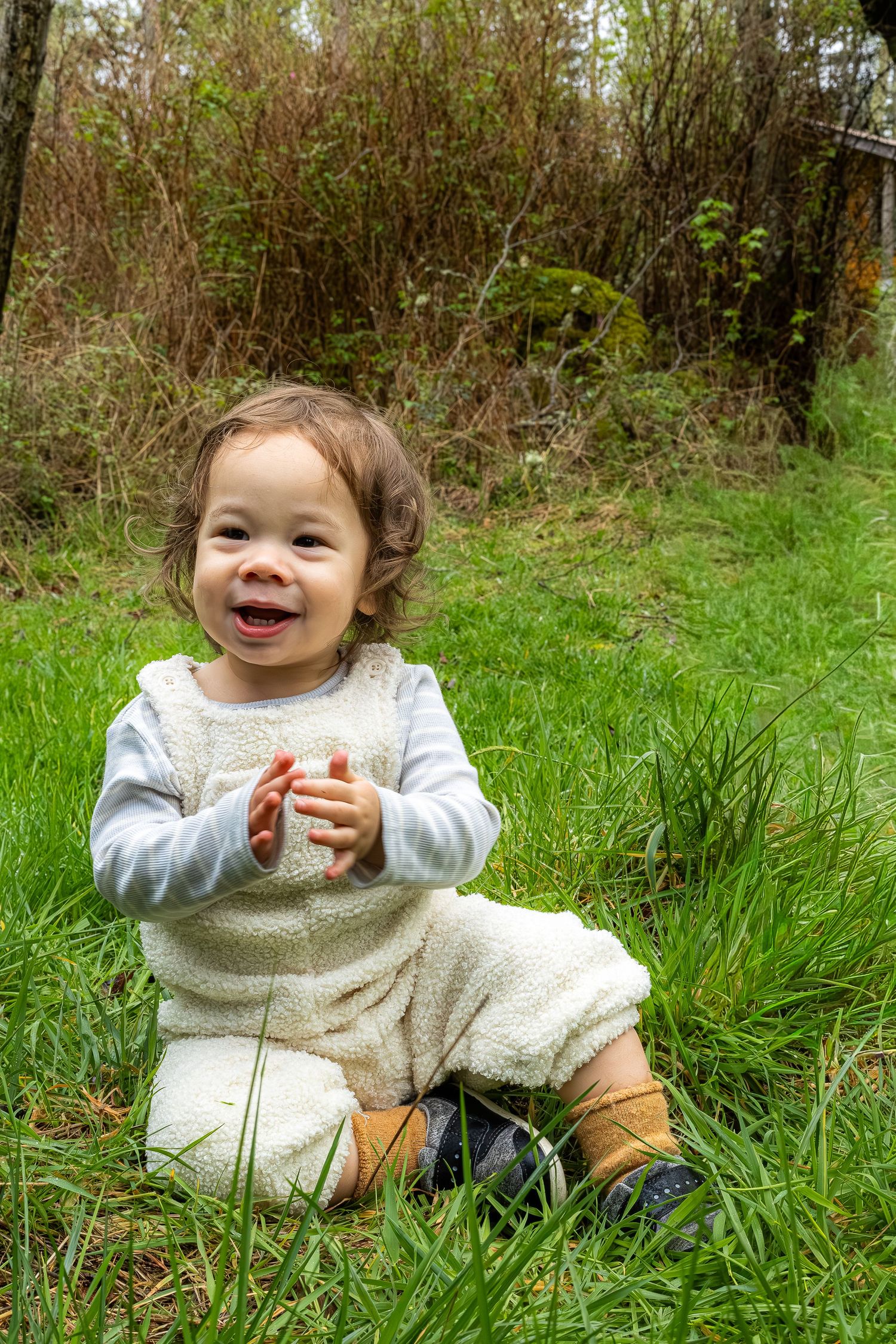 A cheerful baby in a white outfit sits in green grass laughing and clapping hands outdoors.