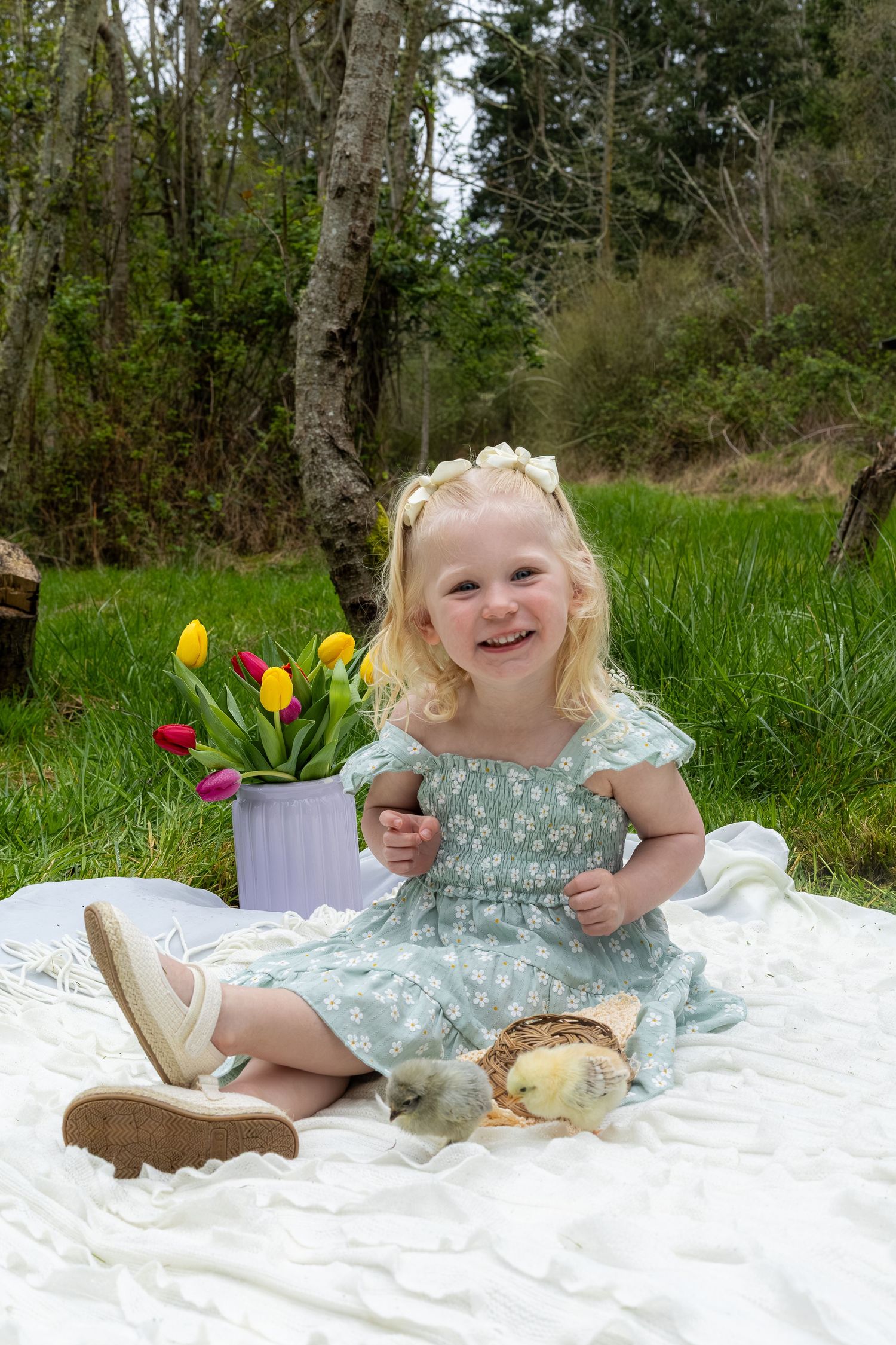 A smiling toddler girl in a blue dress sits on a white blanket with baby chicks and tulips.