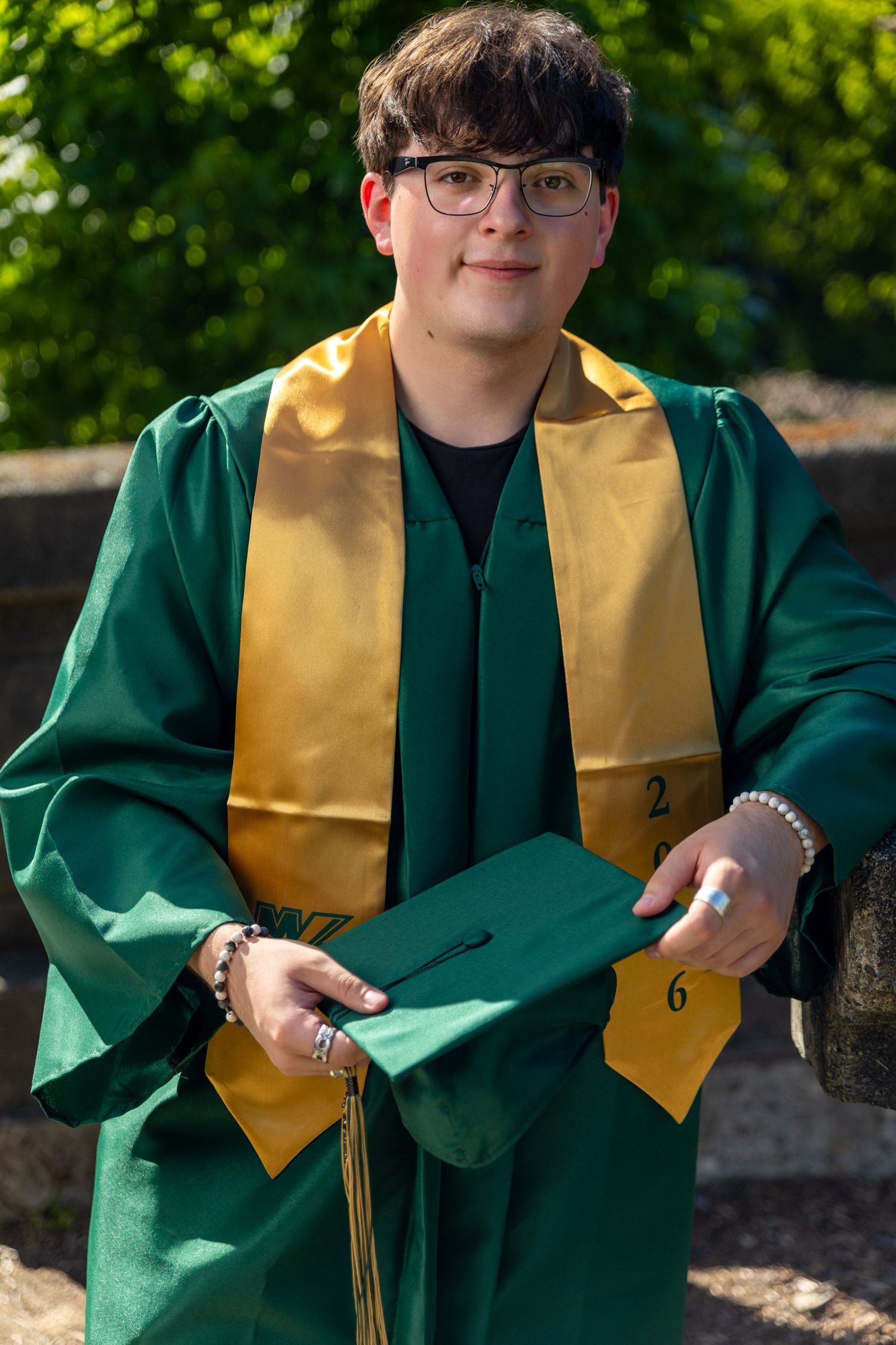 Young graduate in green and gold cap and gown smiling outdoors, holding graduation cap with tassel.