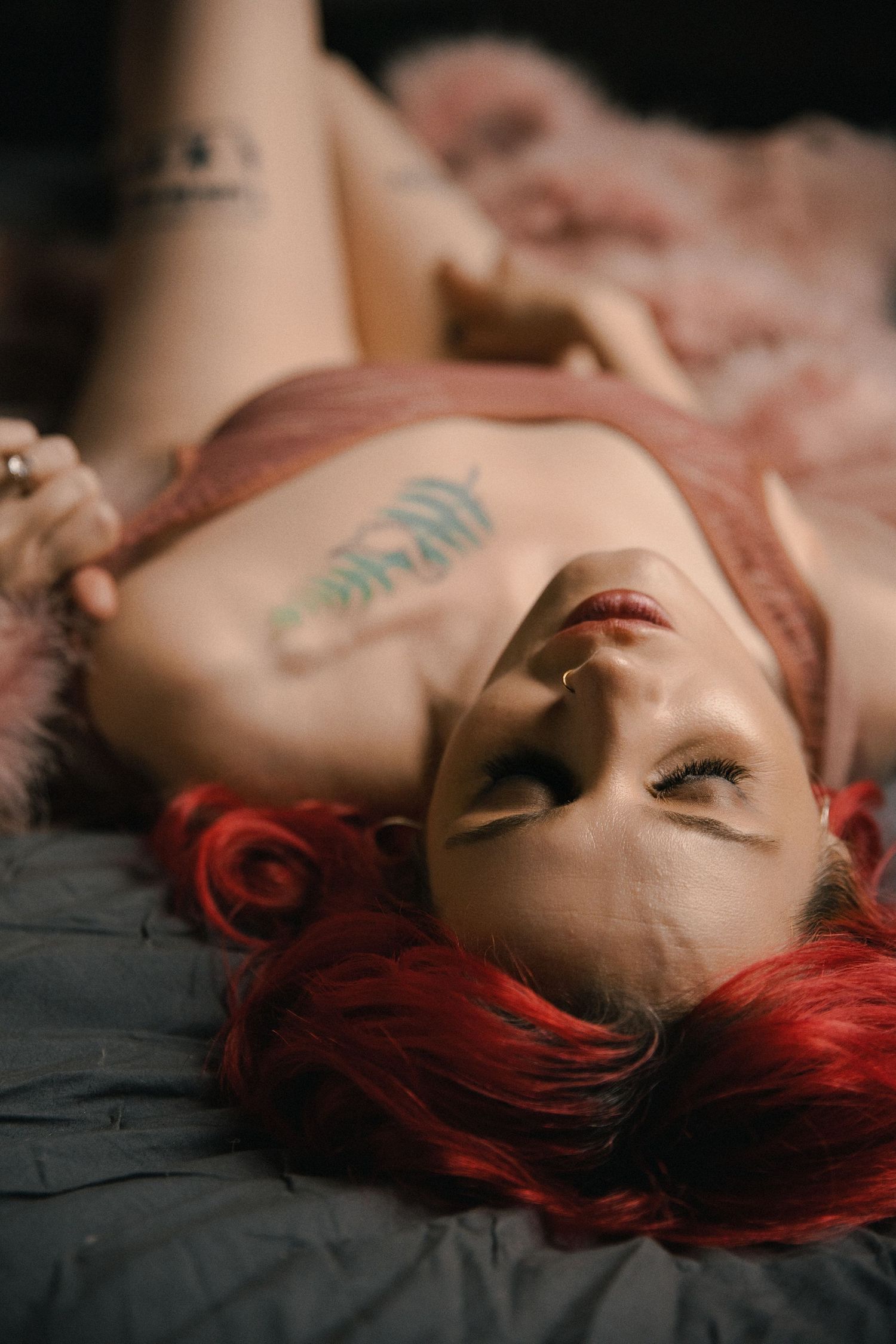 Woman with red hair and tattoos lying on a bed wearing pink lingerie in a boudoir photography session.