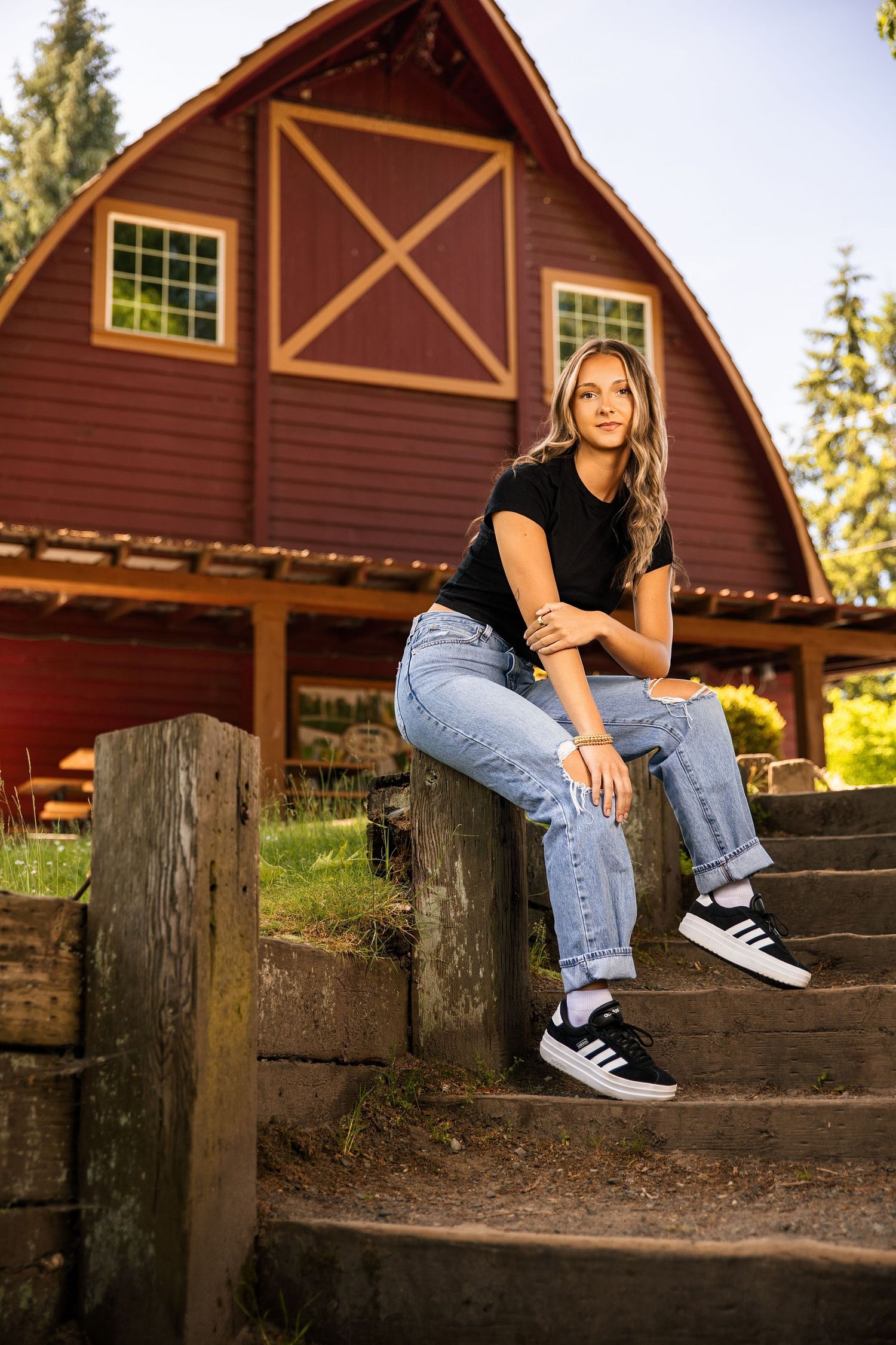 Young woman in black tee and jeans sits on wooden steps before a red barn in golden hour light.