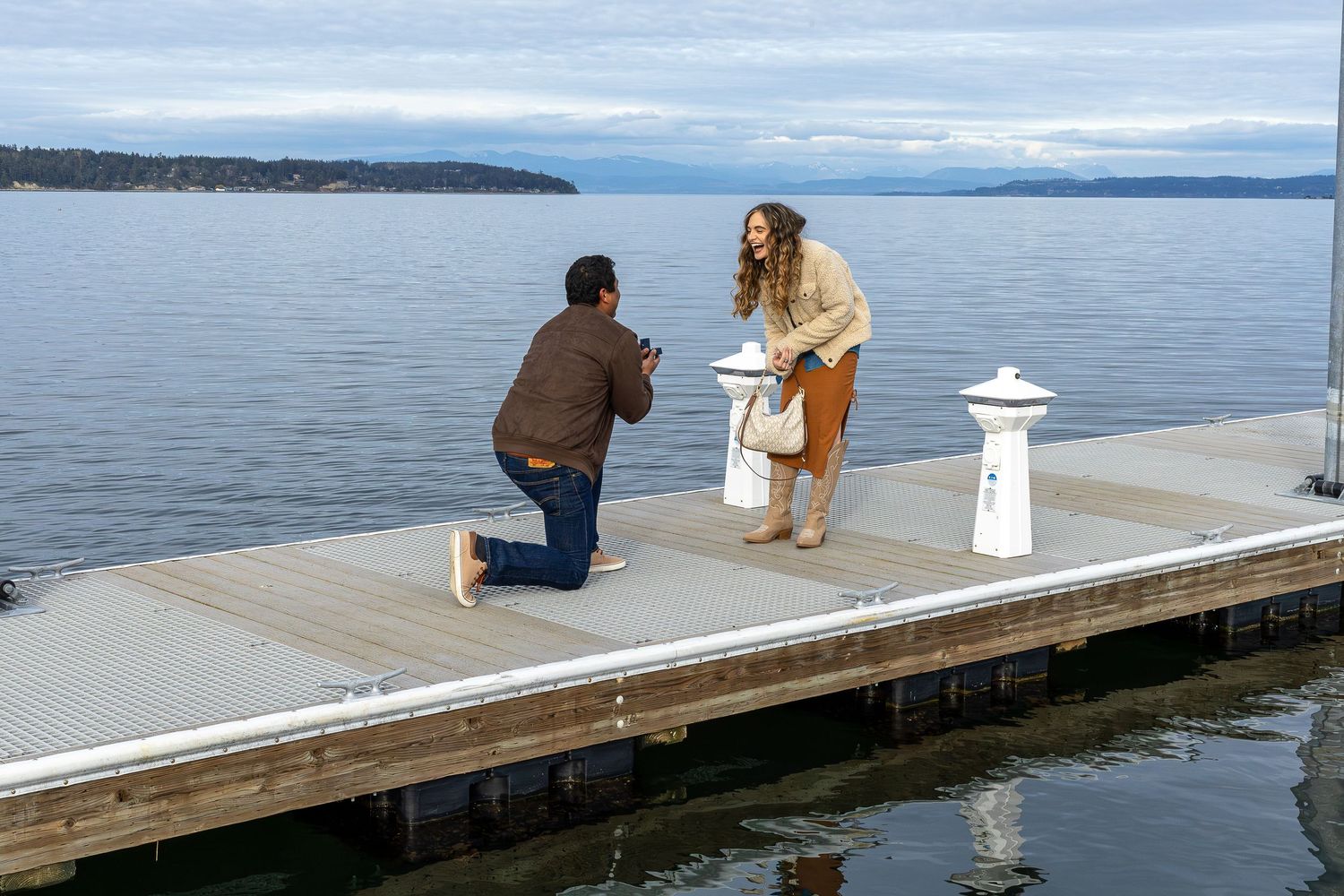 Man proposes to woman on a wooden dock over calm lake water with mountain views in the background.