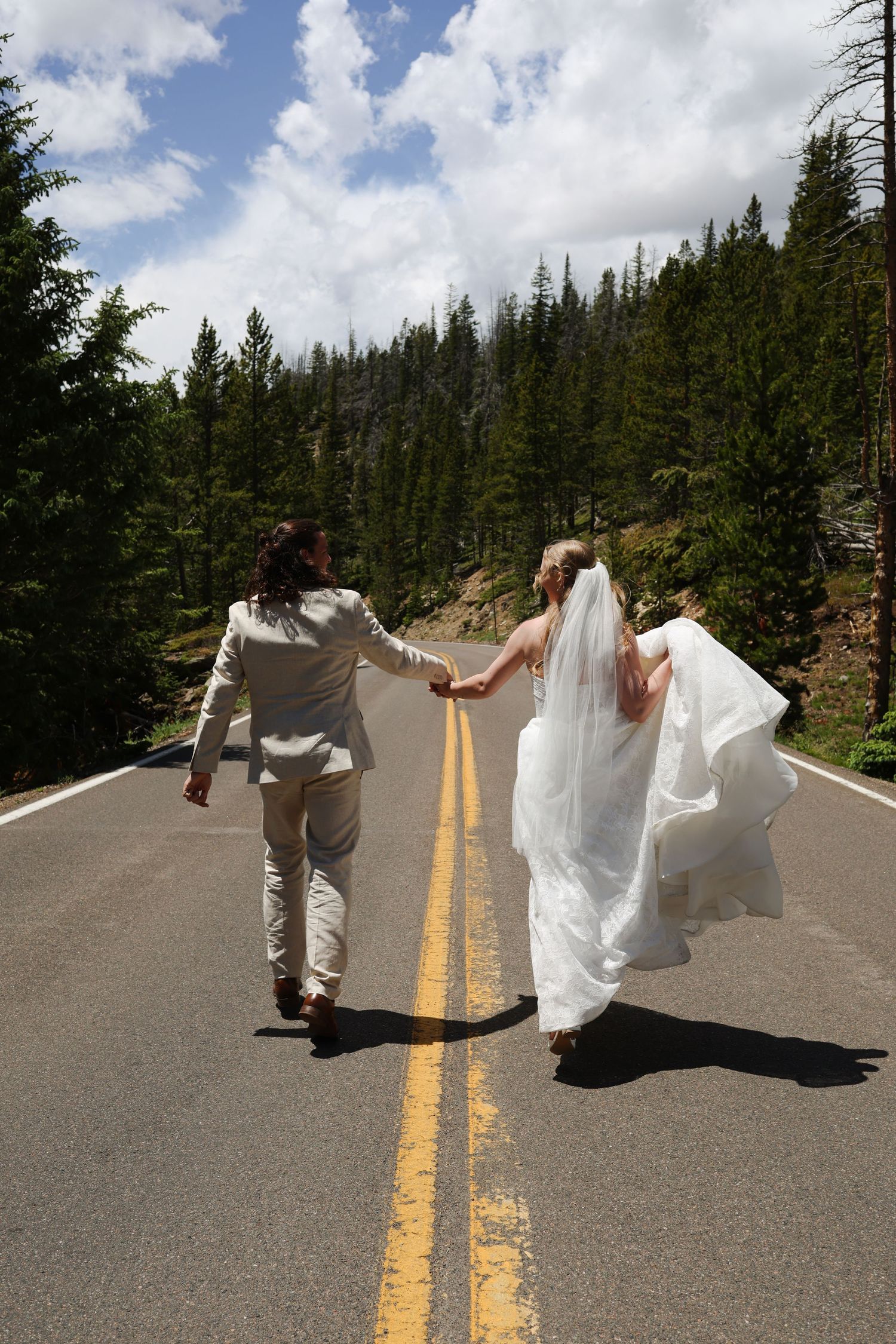 Newlywed couple walks hand-in-hand down a mountain road surrounded by pine trees in wedding attire.