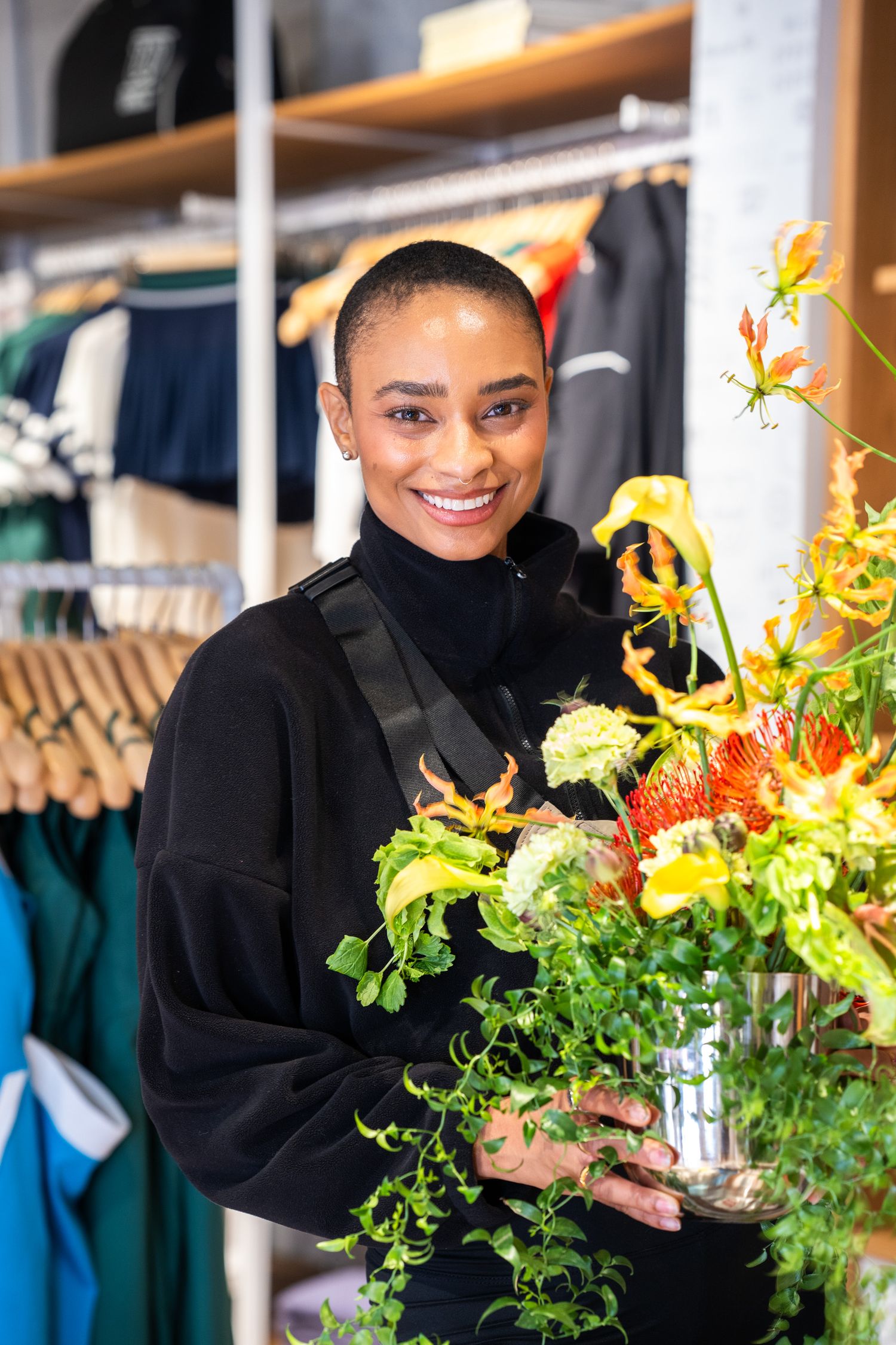 Smiling woman in black top holding vibrant floral arrangement with orange and yellow flowers in a retail store.