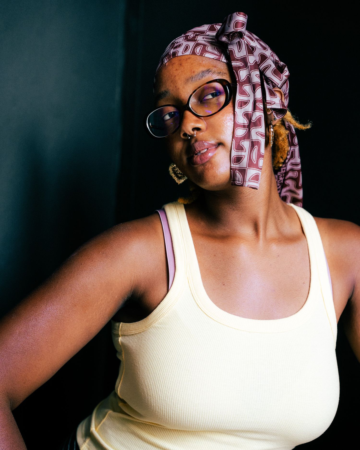 Black woman wearing glasses and a patterned head wrap poses confidently in a cream tank top against dark background.