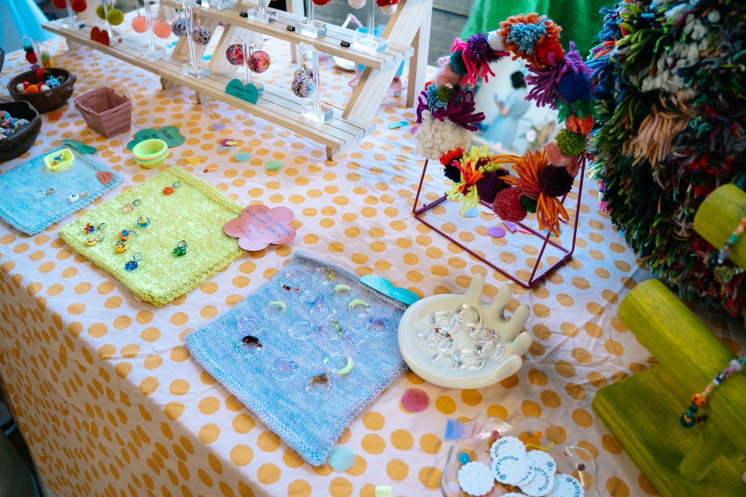 Colorful craft fair table display with handmade jewelry, fabric squares, ceramic bowls, and floral arrangements on polka dot cloth.