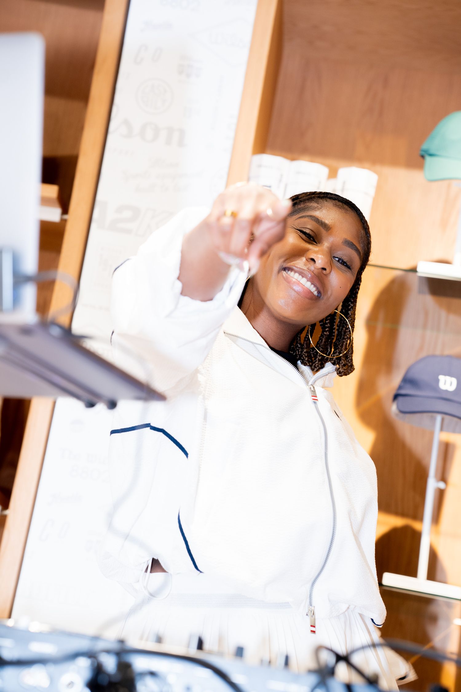 Smiling young Black woman in white jacket reaching toward camera while DJing in a modern retail store.