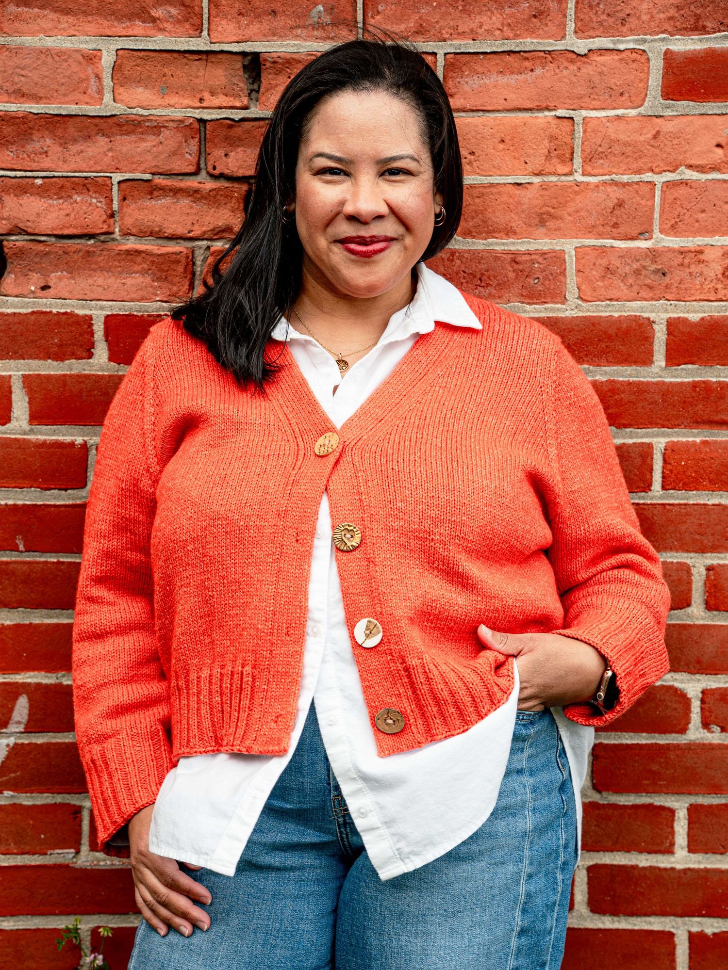 Woman in orange cardigan and jeans smiling against a red brick wall outdoors.
