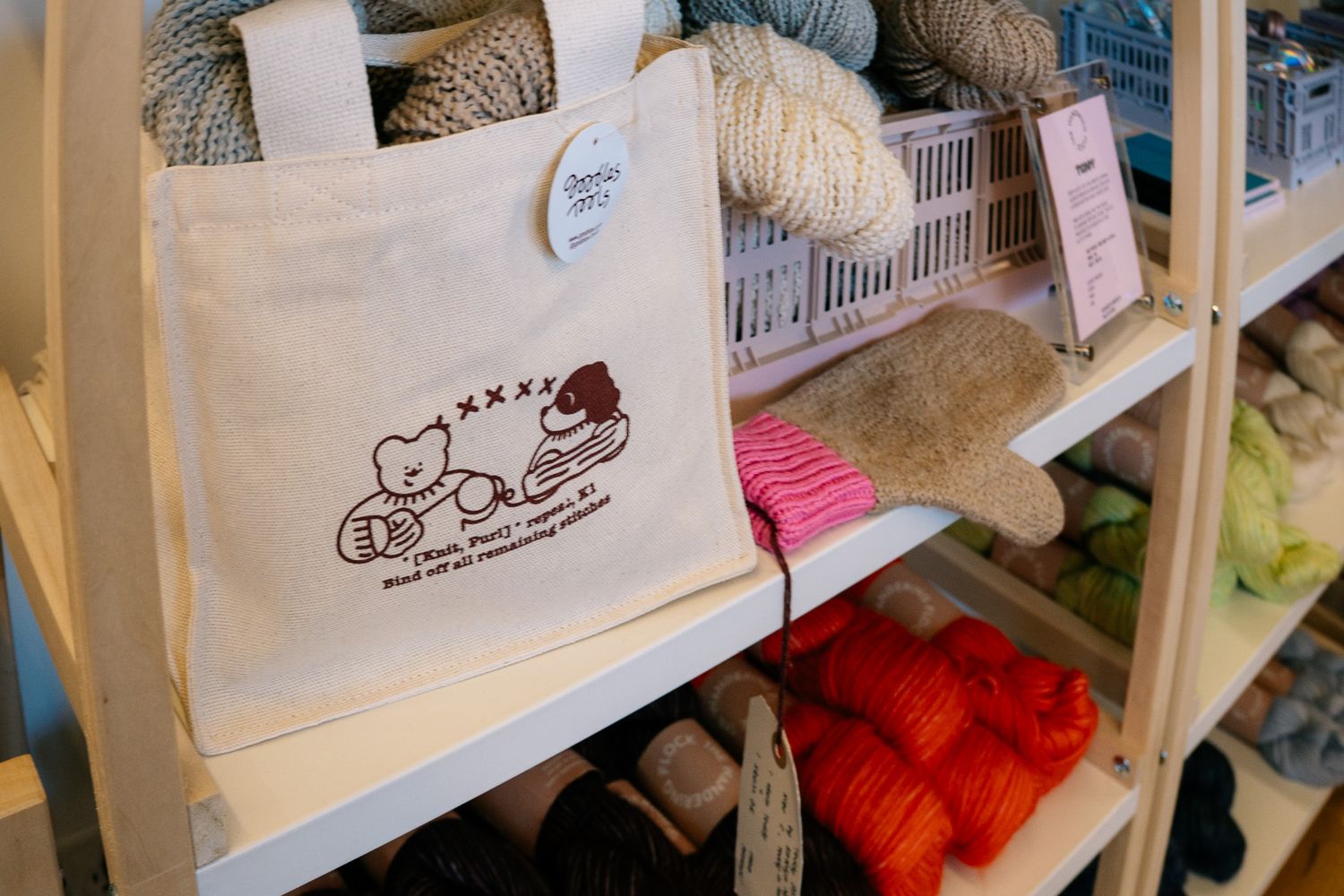 Canvas tote bag with cute bear embroidery displayed on a shelf stocked with colorful yarn skeins in a craft store.