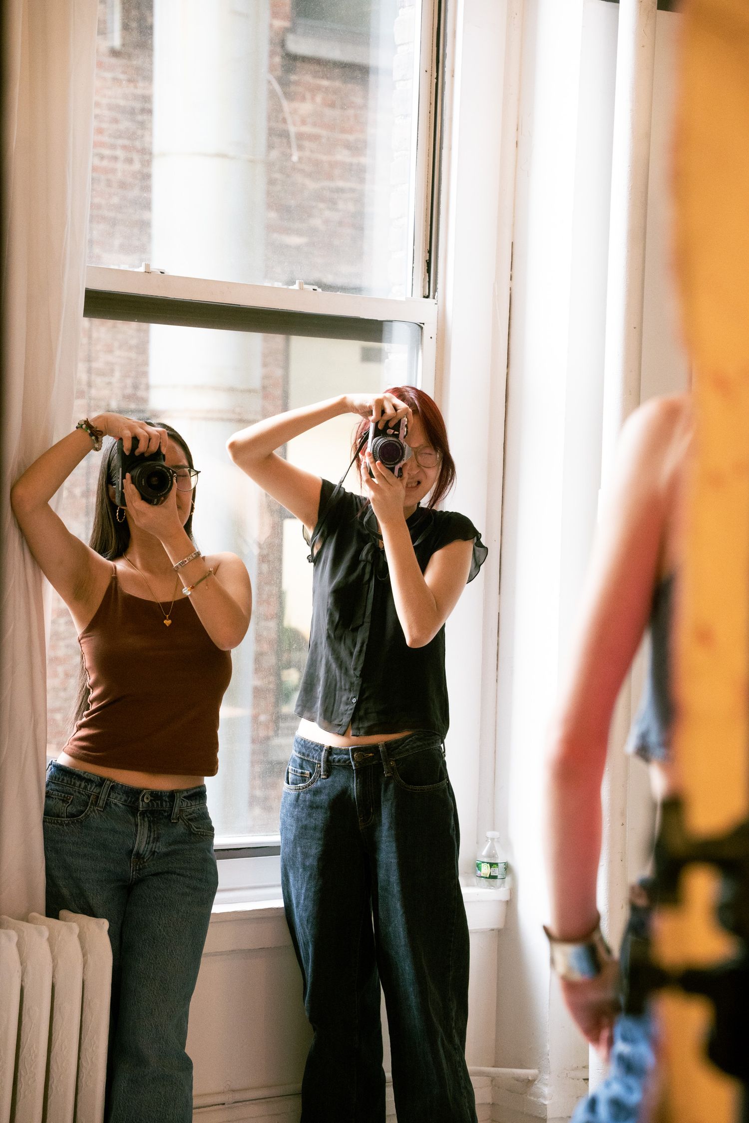 Two women photographing in a sunlit room with cameras raised, wearing casual jeans and tops near large windows.
