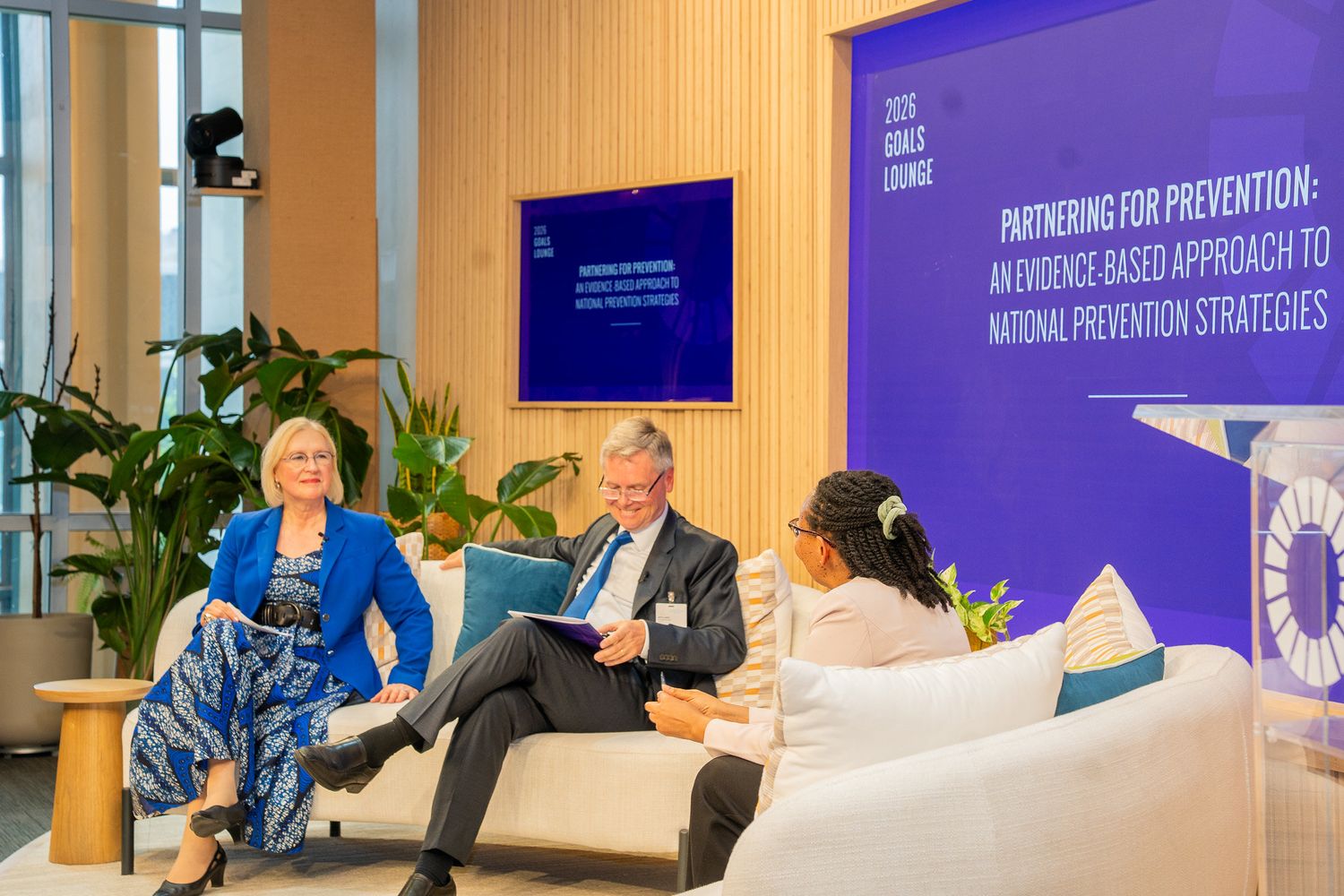 Three professionals in discussion on white sofas at a launch event in the United Nations Headquarters SDG Lounge.