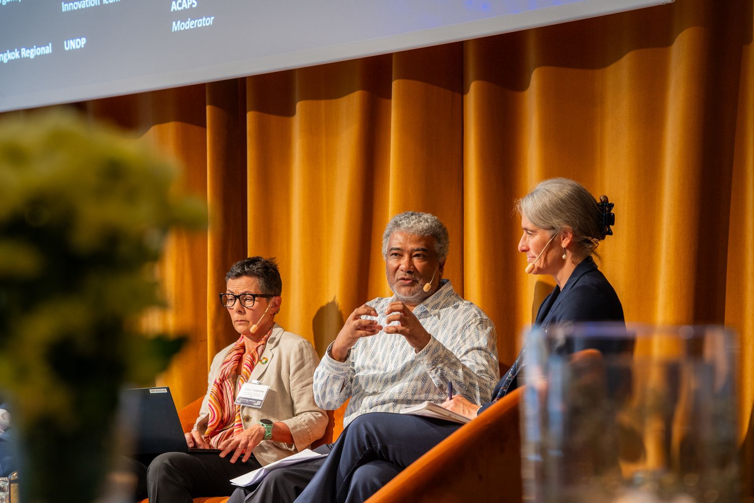 Three panelists engaged in discussion on stage with orange curtain backdrop at a conference.