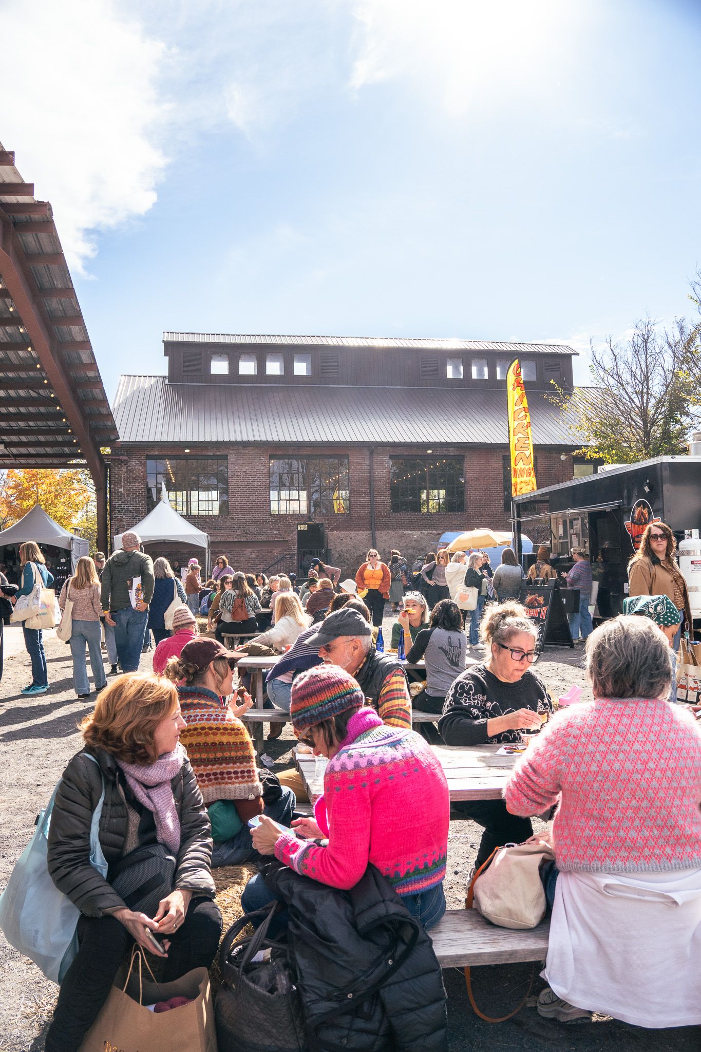 Crowds gather at an outdoor market near a brick building on a sunny autumn day, with people sitting at picnic tables.