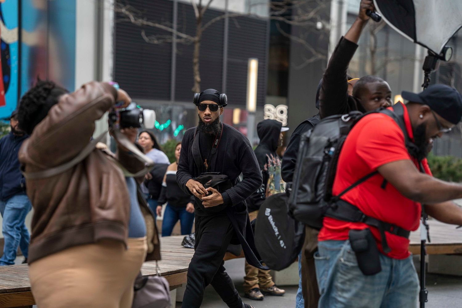 Group of people on a busy urban street, with a man in black outfit walking confidently past photographers.