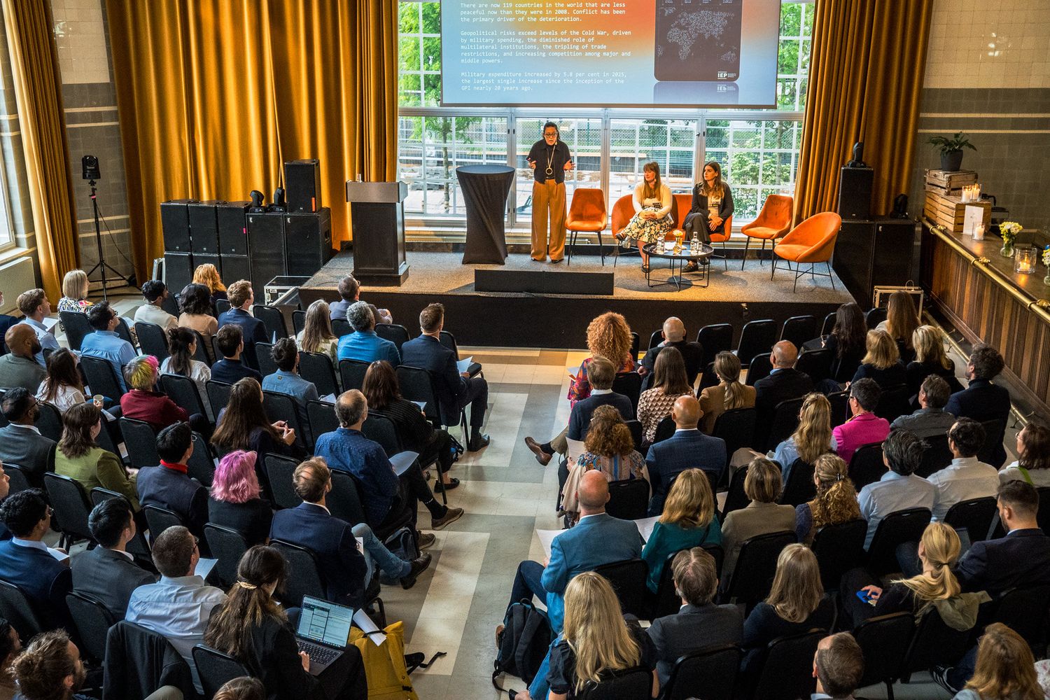 Large audience attending a conference presentation in an elegant auditorium with a lit stage.