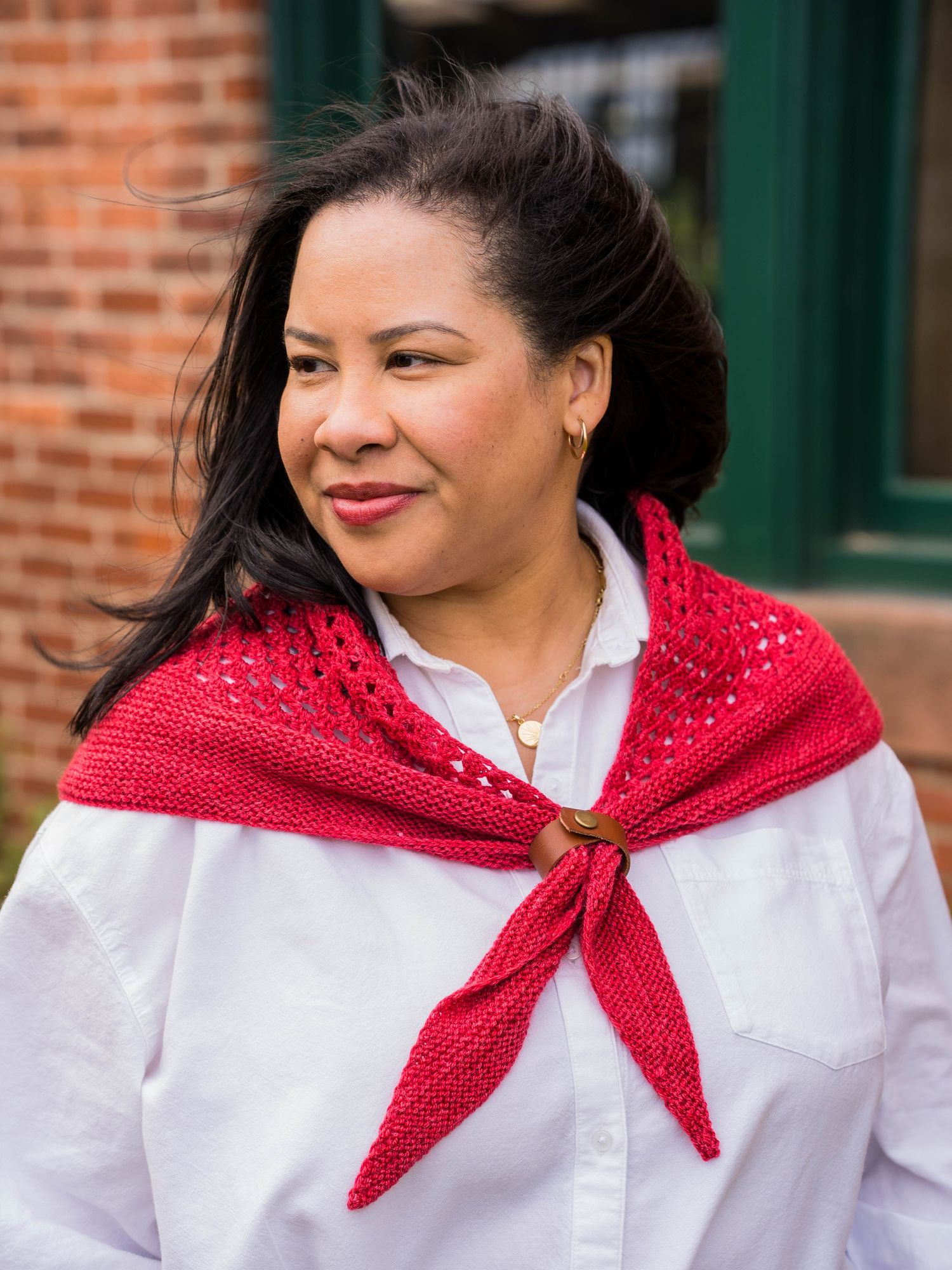 Woman wearing a red knitted shawl over a white shirt, standing outdoors near a brick building.