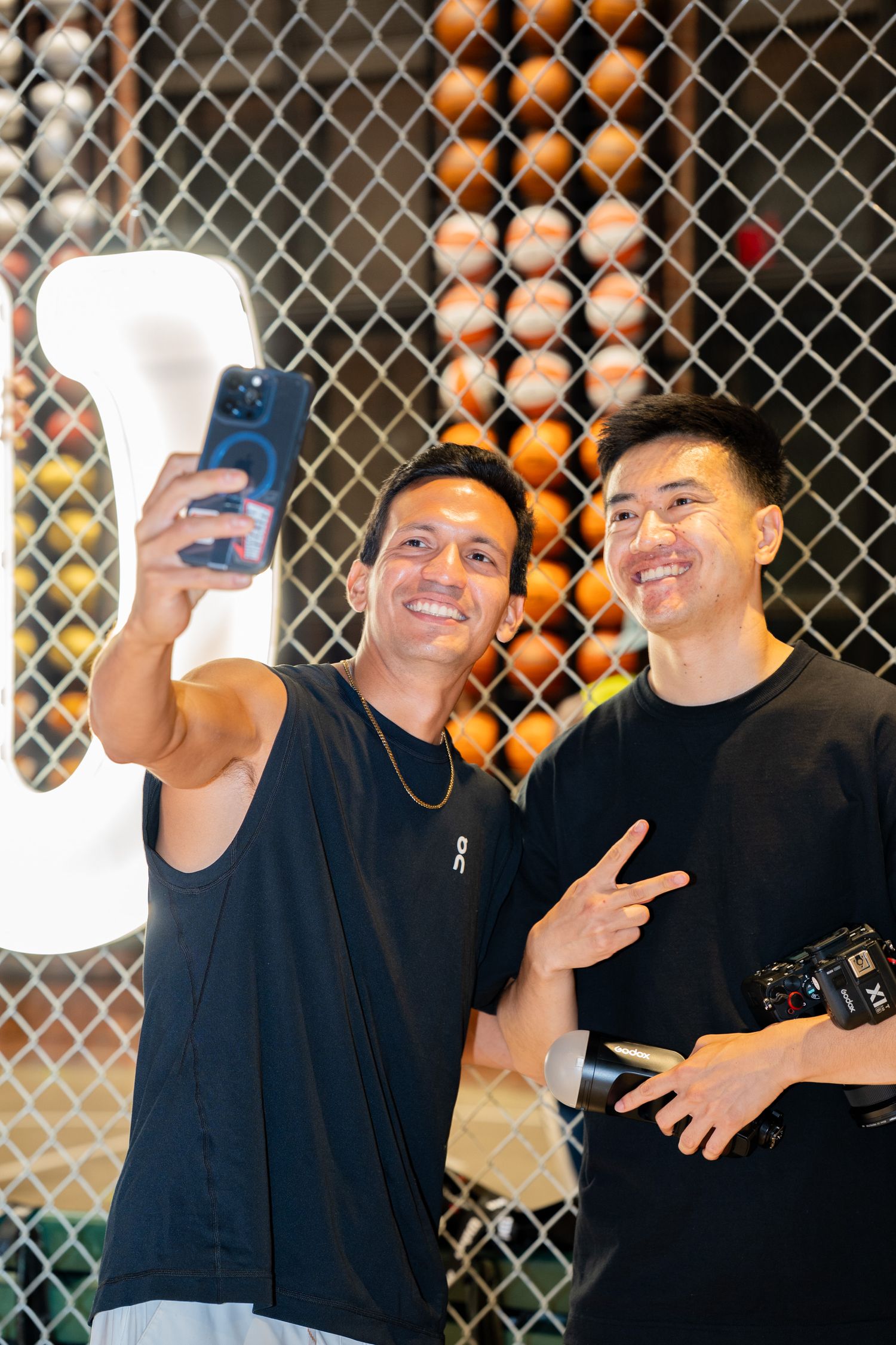 Two smiling men in black shirts take a selfie together, one holding a camera, in front of a chain-link fence backdrop.