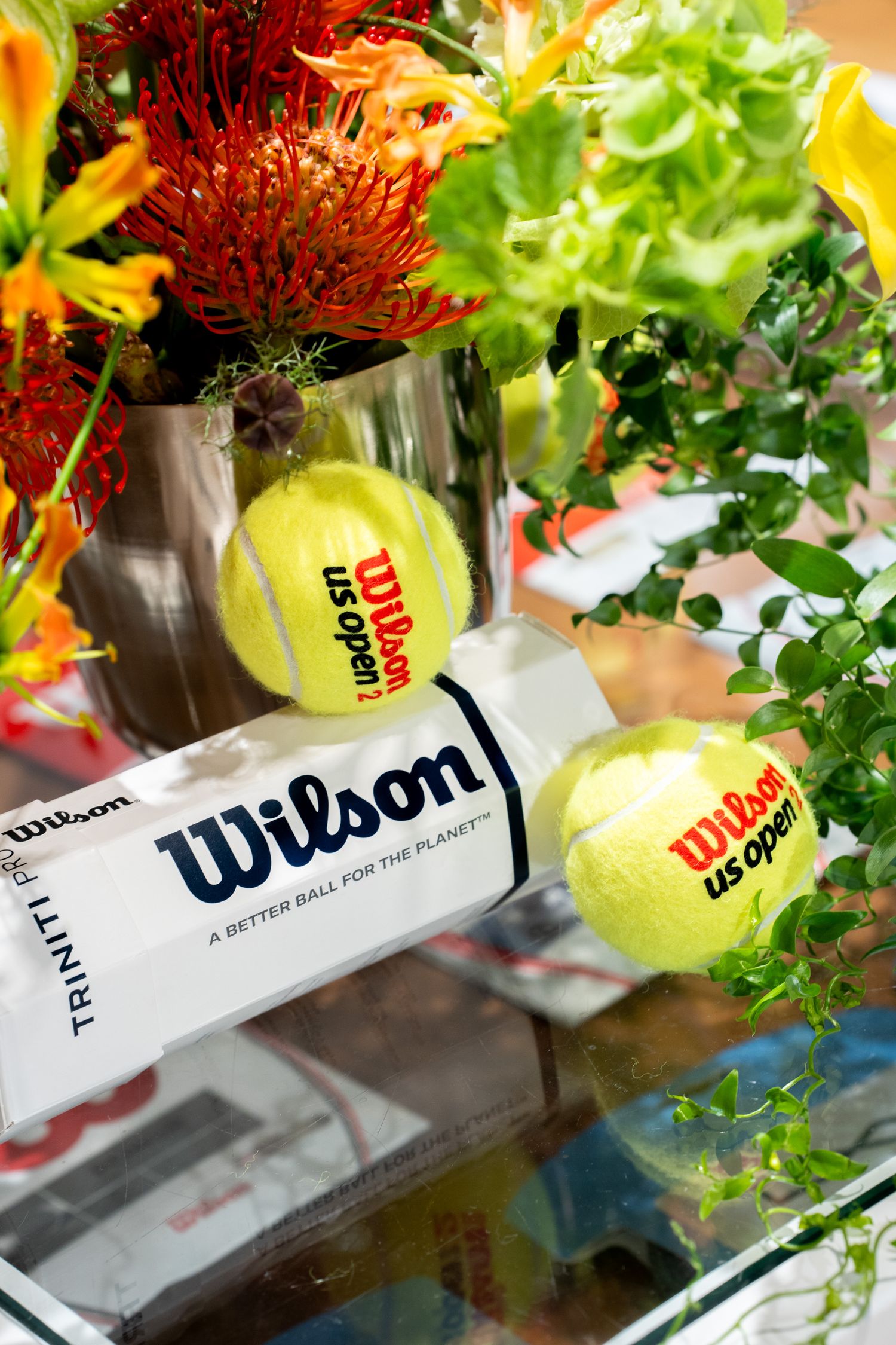 Wilson US Open tennis balls displayed beside a vibrant floral arrangement in a silver vase on a glass table.