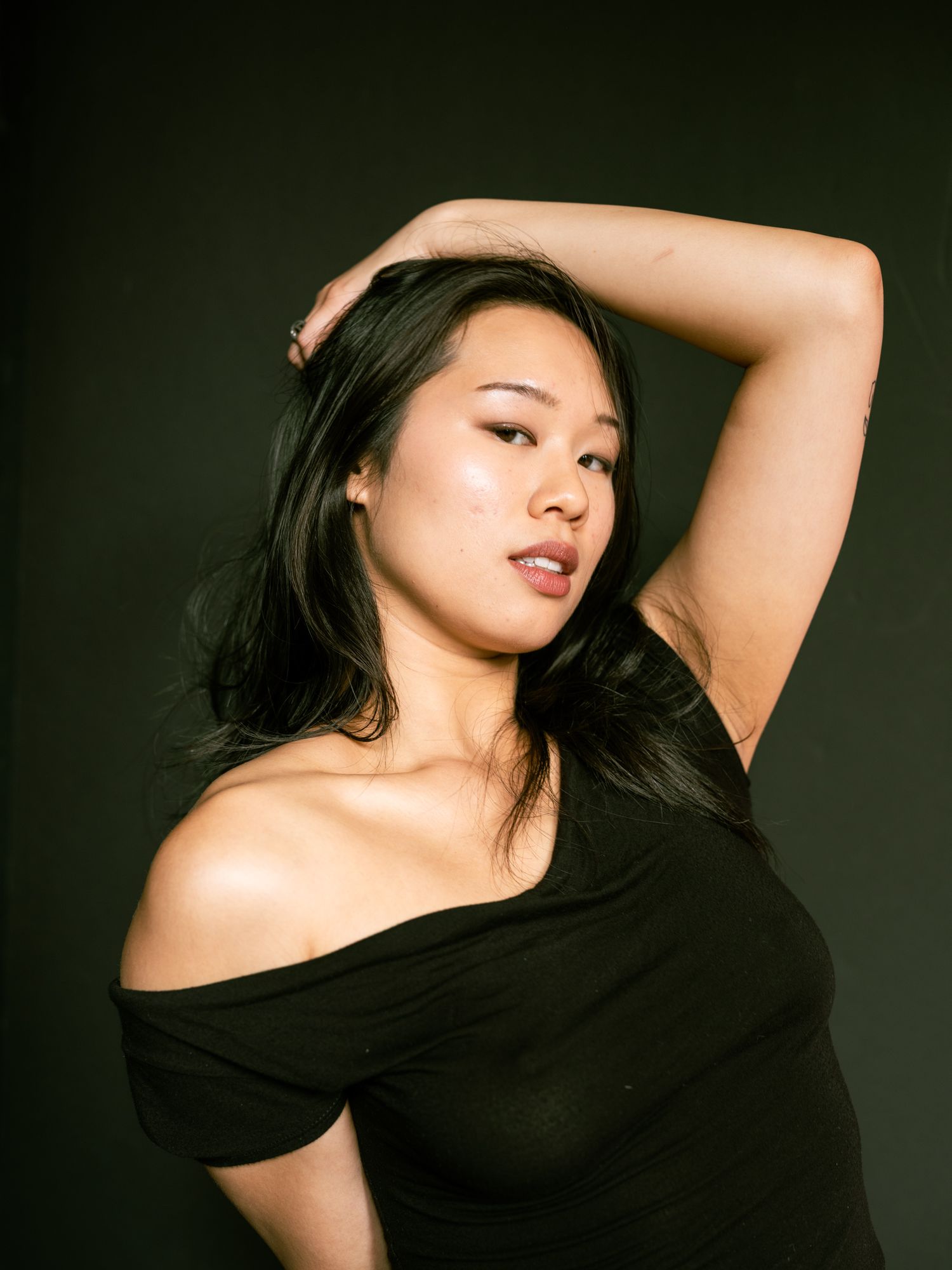 An Asian woman in a black off-shoulder top poses gracefully with one hand raised to her head against a dark background.
