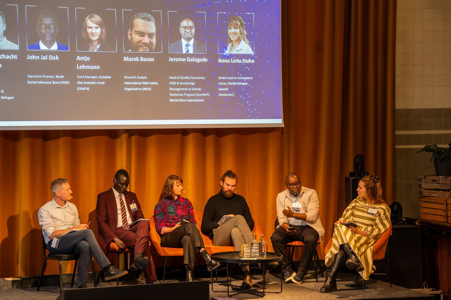 Seven diverse panelists seated on stage beneath a projected speaker profile screen at a summit.