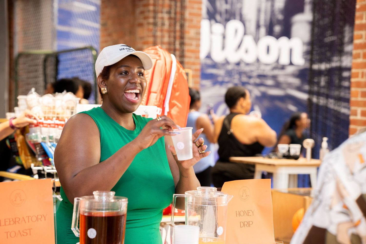 Woman in green dress and white cap smiling at a busy indoor market with drinks and snacks on the table.