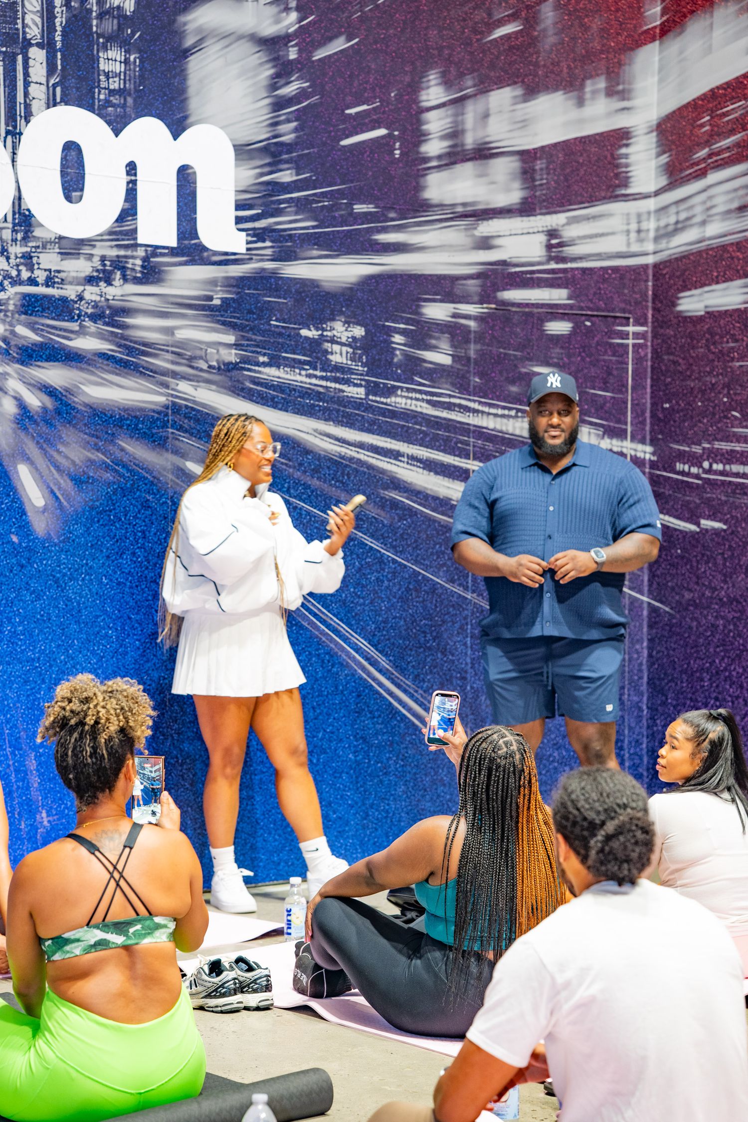 Two presenters speak to a seated audience in front of a blue graphic backdrop at an indoor event.