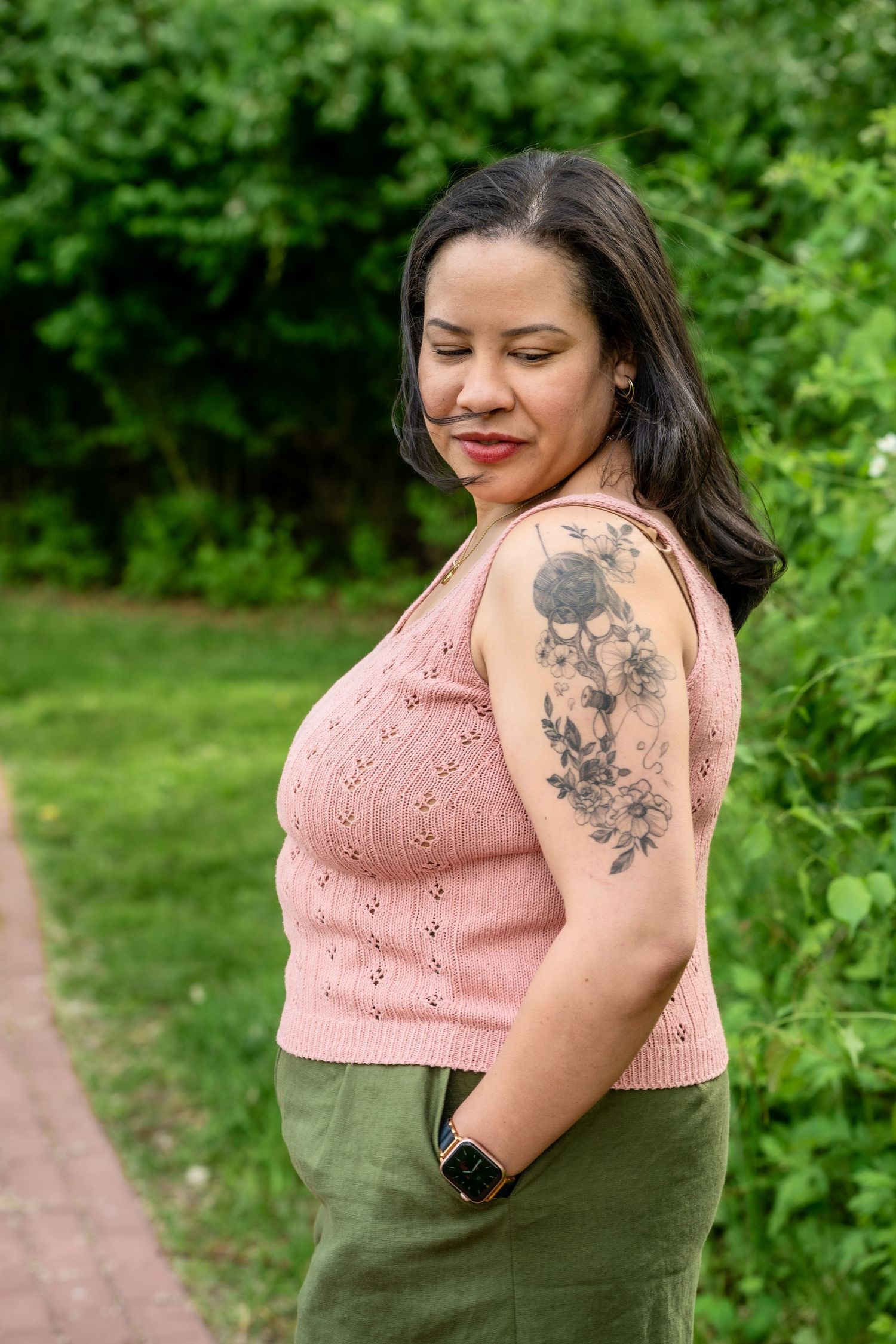 Woman with floral arm tattoo wearing pink knit top and green skirt, posing outdoors among lush greenery.