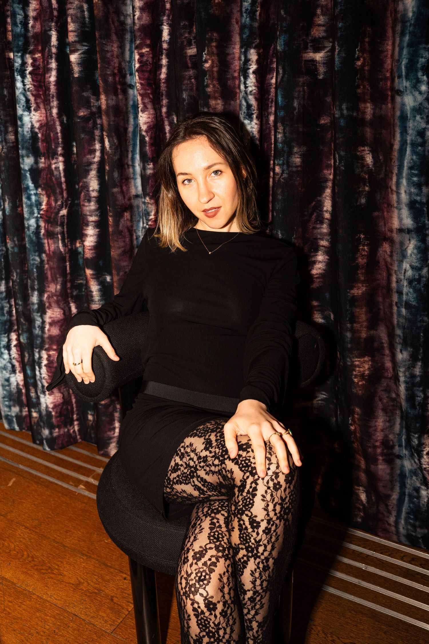 Woman in black dress and lace tights seated in chair against colorful velvet curtain backdrop.