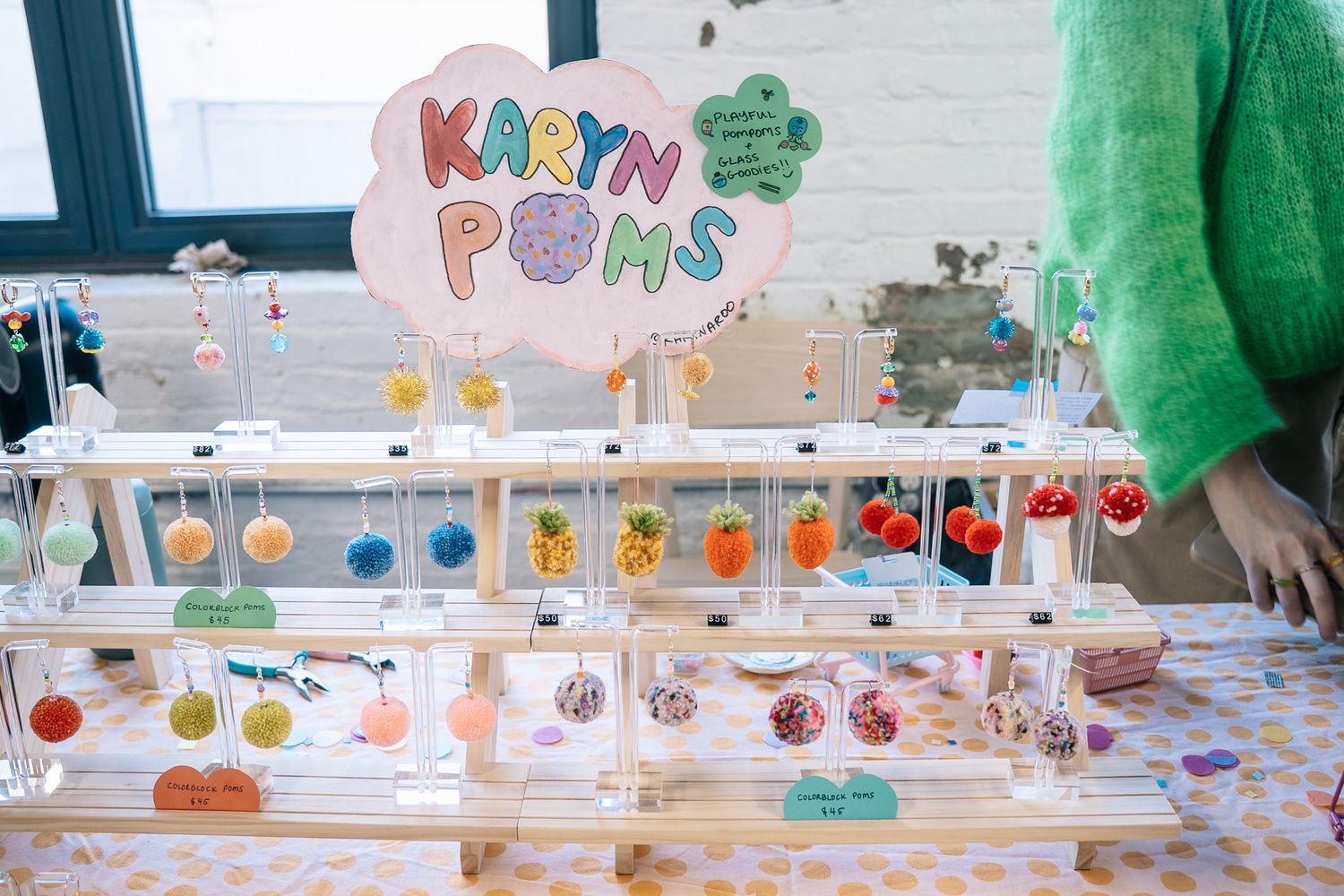 Karyn Poms colorful handmade pom-pom jewelry display on tiered acrylic stand at a craft fair.