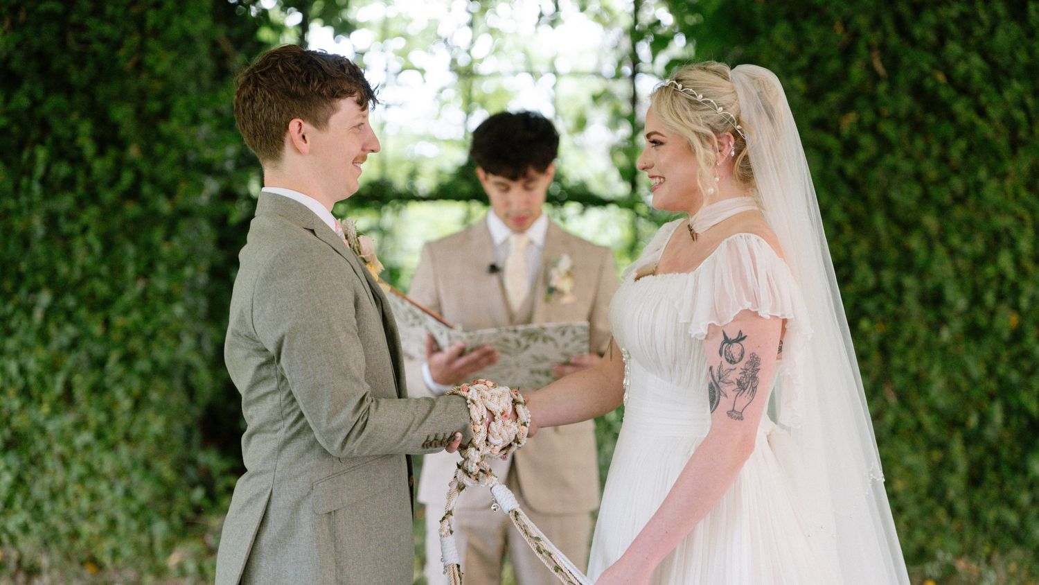 Bride and groom exchange vows outdoors beneath lush green ivy wall with officiant present.