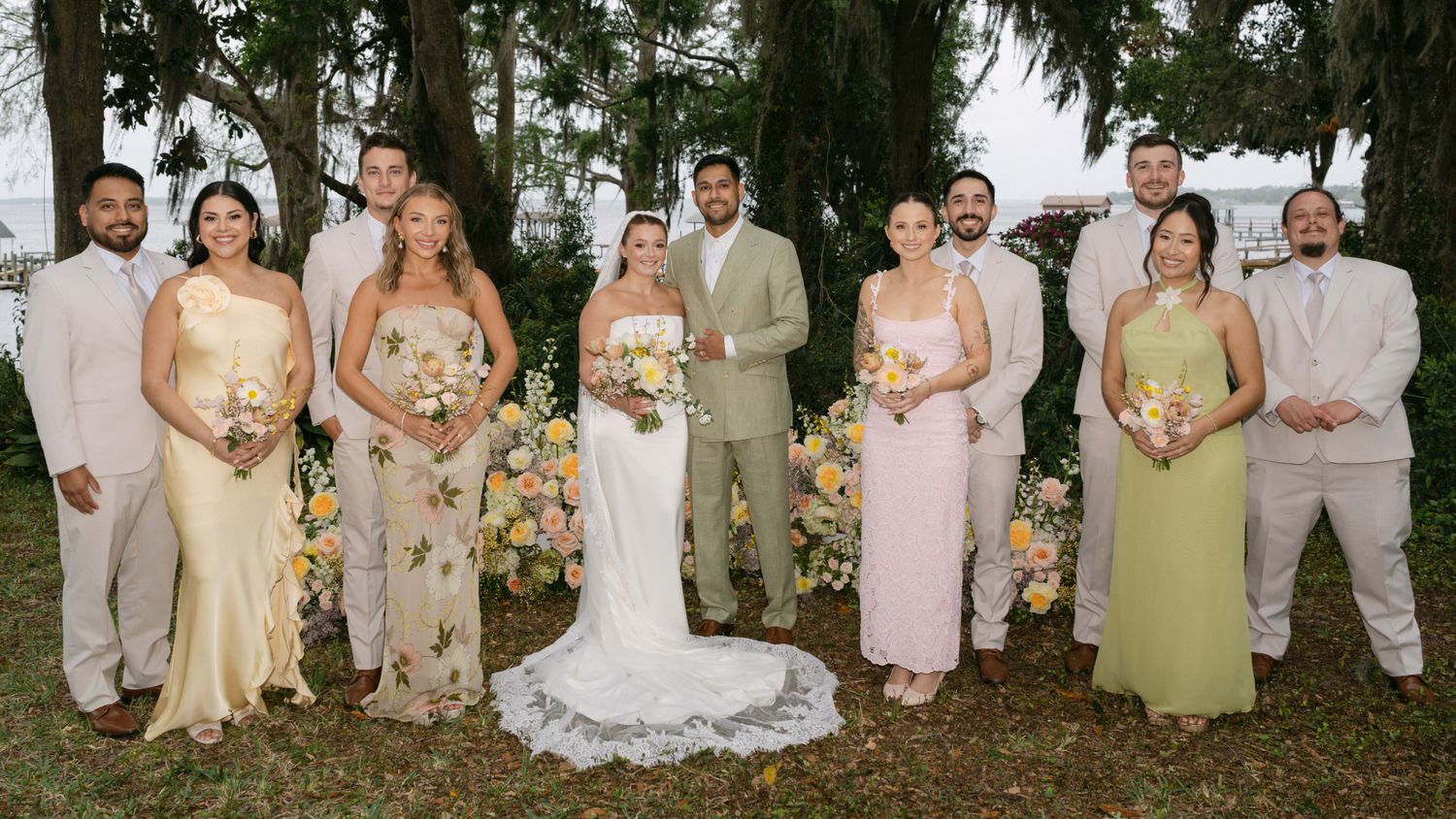 Wedding party poses together outdoors in pastel attire with colorful floral arrangements nearby.