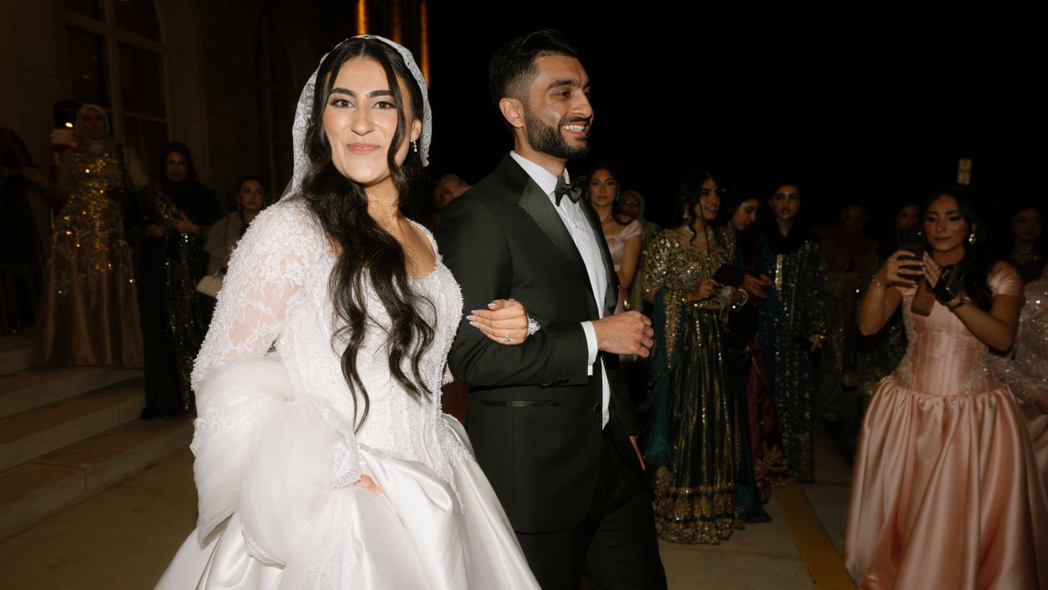 Bride in white gown and groom in tuxedo make grand entrance at elegant evening wedding celebration.