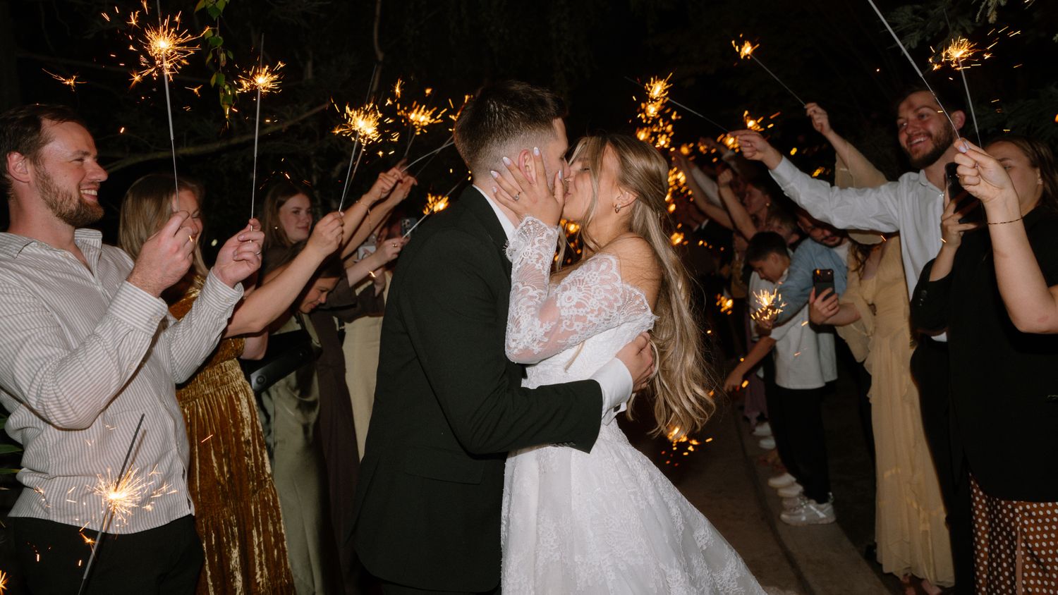 Newlyweds share a kiss surrounded by guests holding sparklers during festive wedding sendoff.