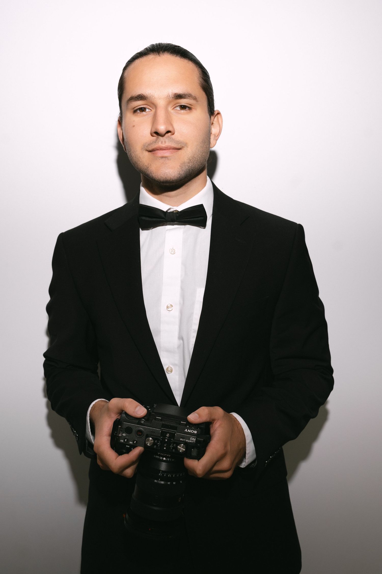 Professional wedding photographer in tuxedo holding camera poses against white background.