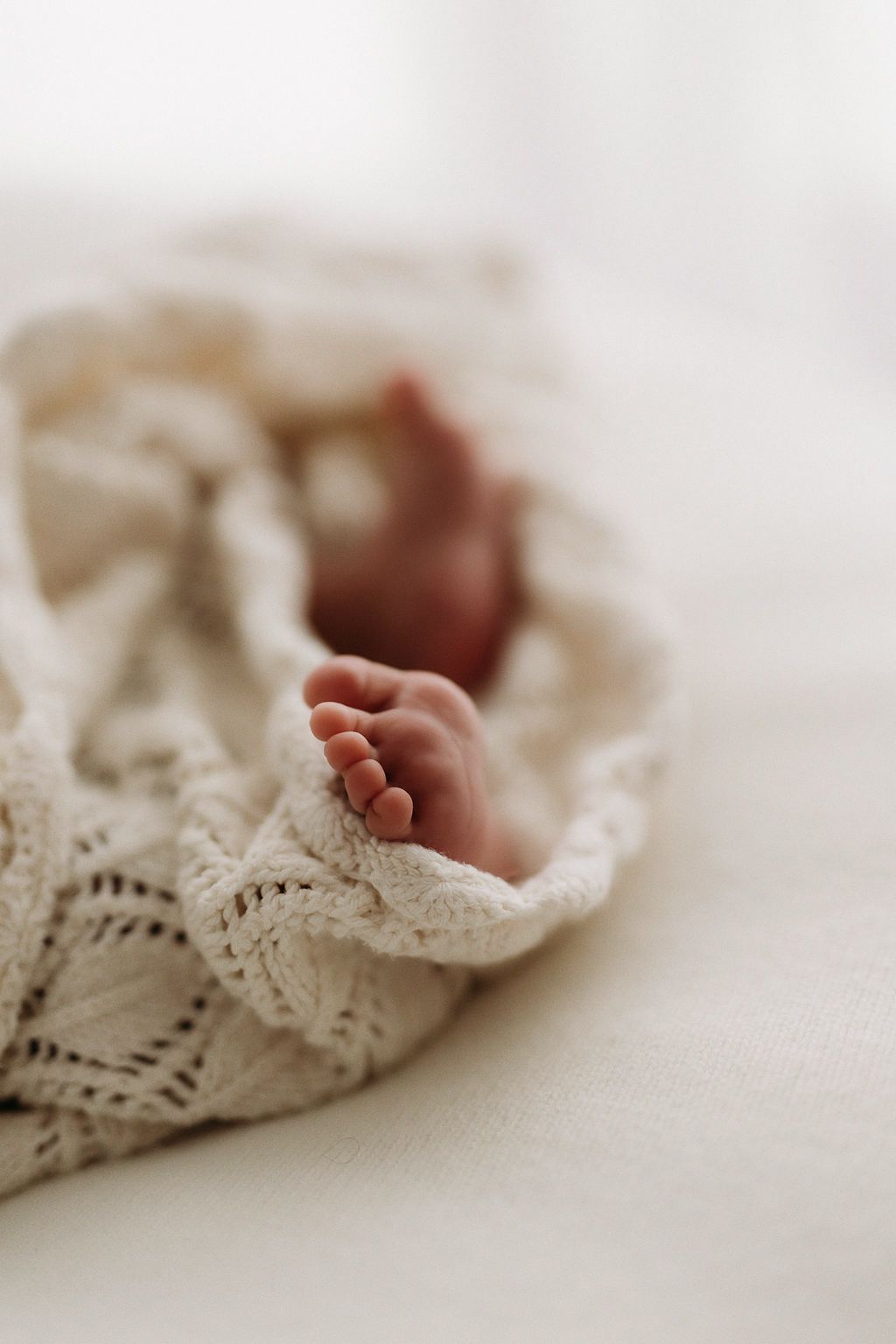 Tiny newborn baby feet peeking out from a soft cream knit lace blanket on a white surface.
